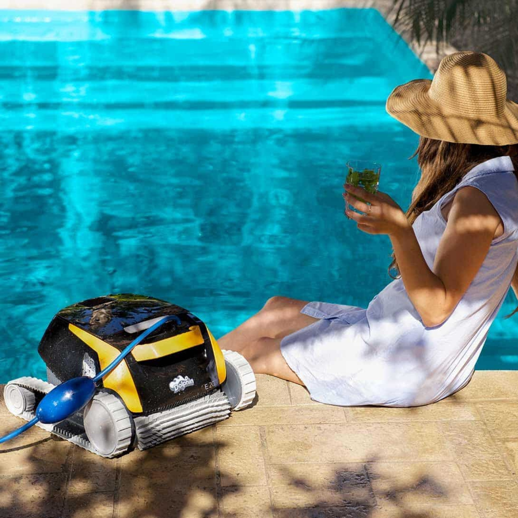 Una mujer con un vestido blanco y sombrero se relaja junto a una piscina, con un limpiador y una bebida cerca.