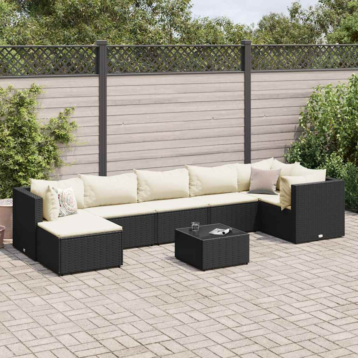 Ein schwarzes Korbsofa mit cremefarbenen Kissen und ein schwarzer quadratischer Tisch auf einer Terrasse. Ein Holzzaun steht.