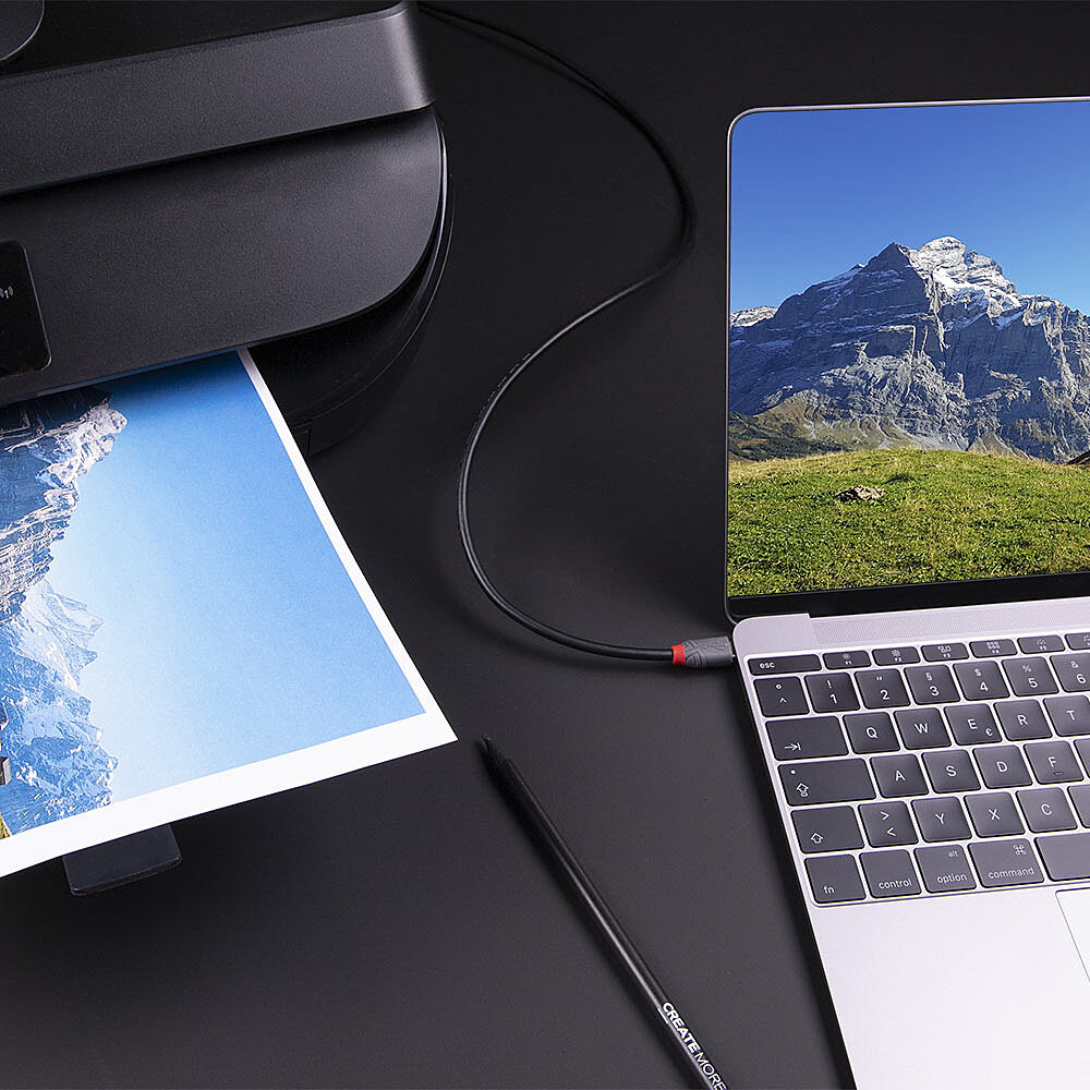 Laptop druckt ein Bild von einem Drucker. Ein Stift liegt auf dem Tisch. Der Bildschirm zeigt einen Berg.