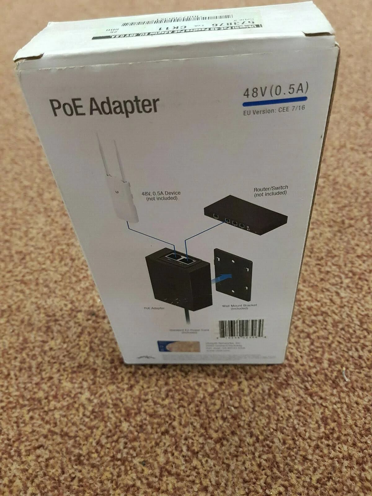 Eine weiße Box mit Text und einem Diagramm, das einen PoE-Adapter und zugehörige Geräte zeigt. Die Box steht auf einem Teppich.