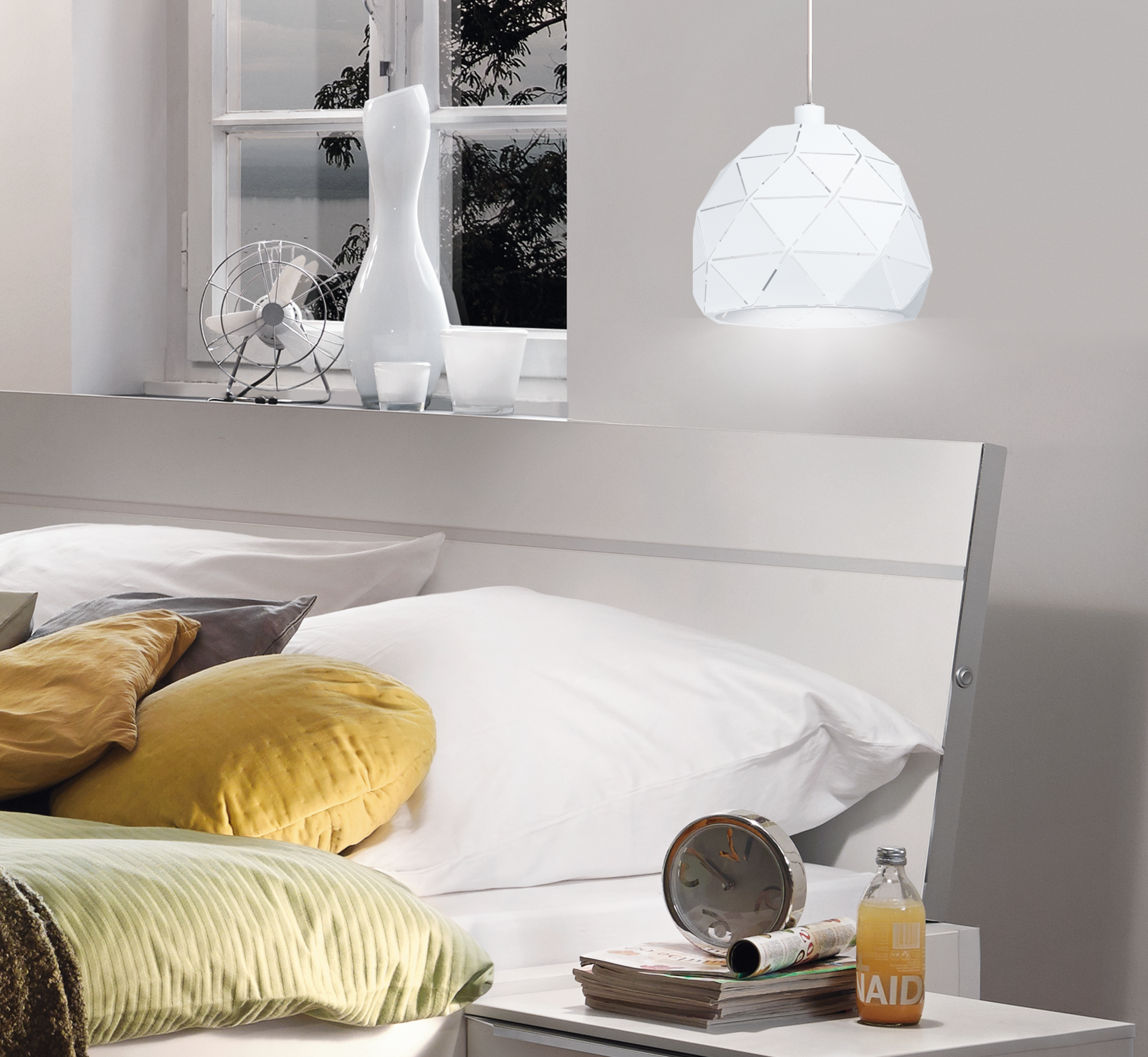 Una escena de dormitorio con cabecero blanco, lámpara blanca, ventilador y mesa con objetos.