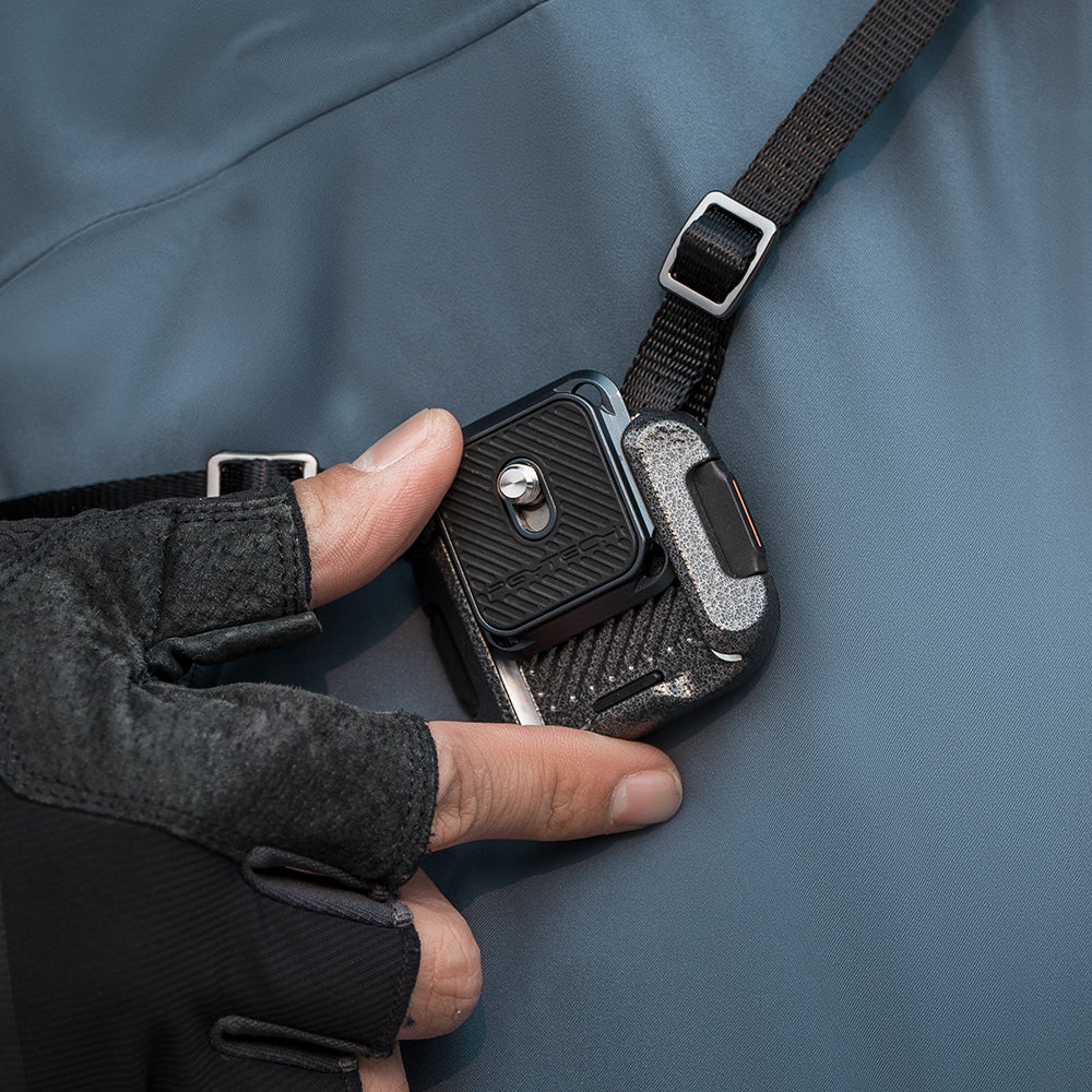 La mano di una persona che tiene un accessorio per fotocamera nero e argento attaccato a un cinturino sull'indumento.