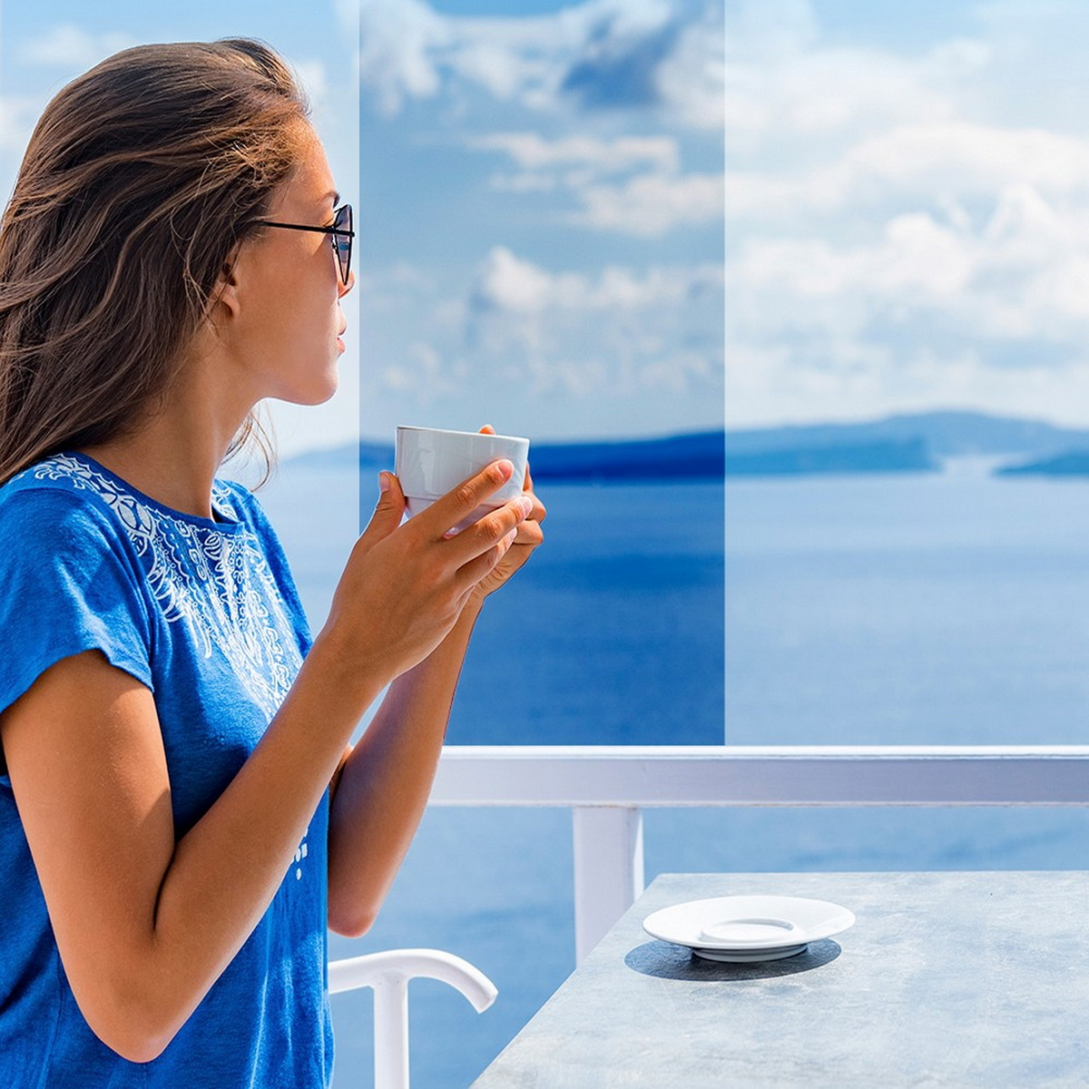 Eine Frau hält eine Tasse und blickt aufs Meer. Ein Tisch und ein Geländer sind im Vordergrund.
