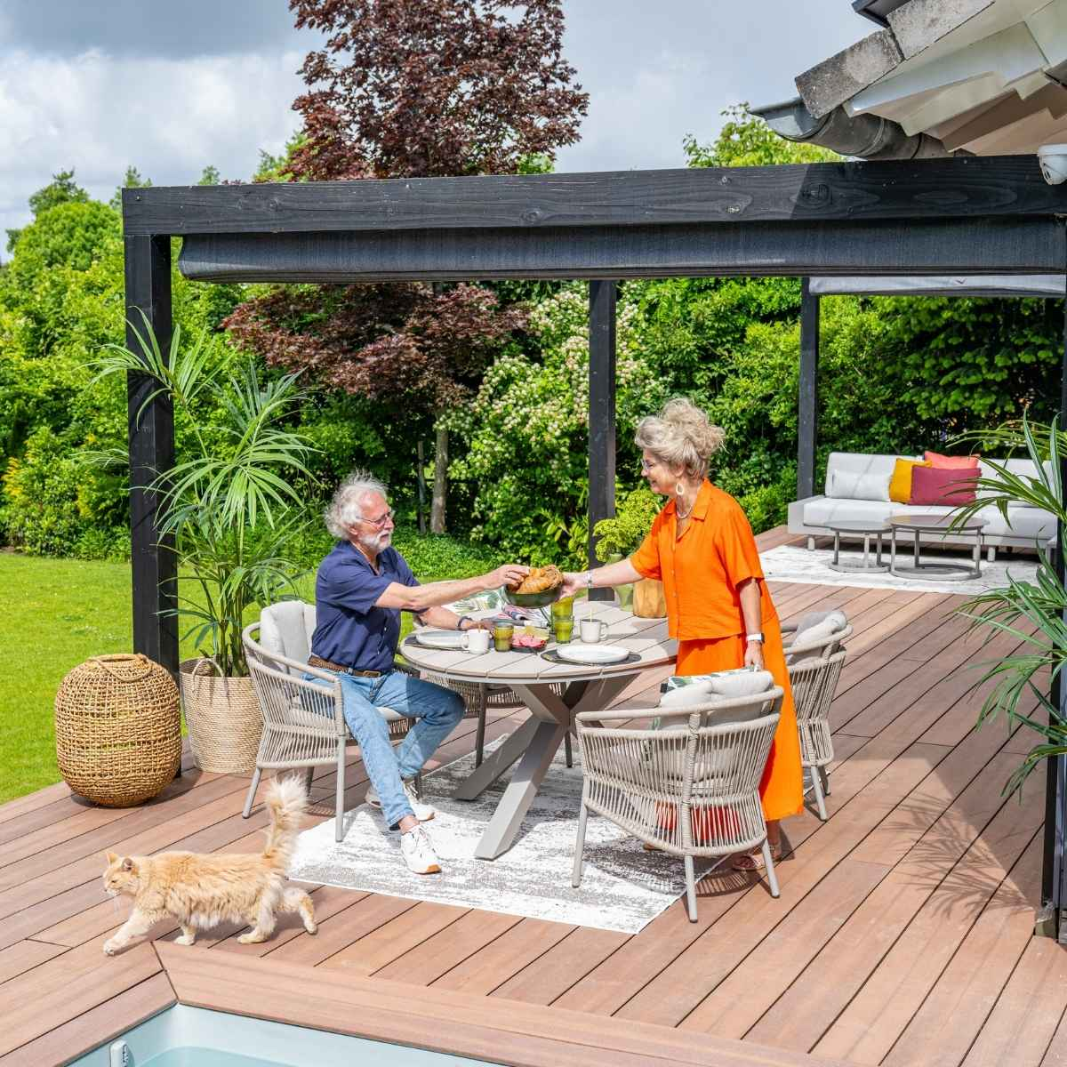 Een stel zit aan een tafel op een terras. Een kat loopt voorbij. Veel groen omringt het terras.