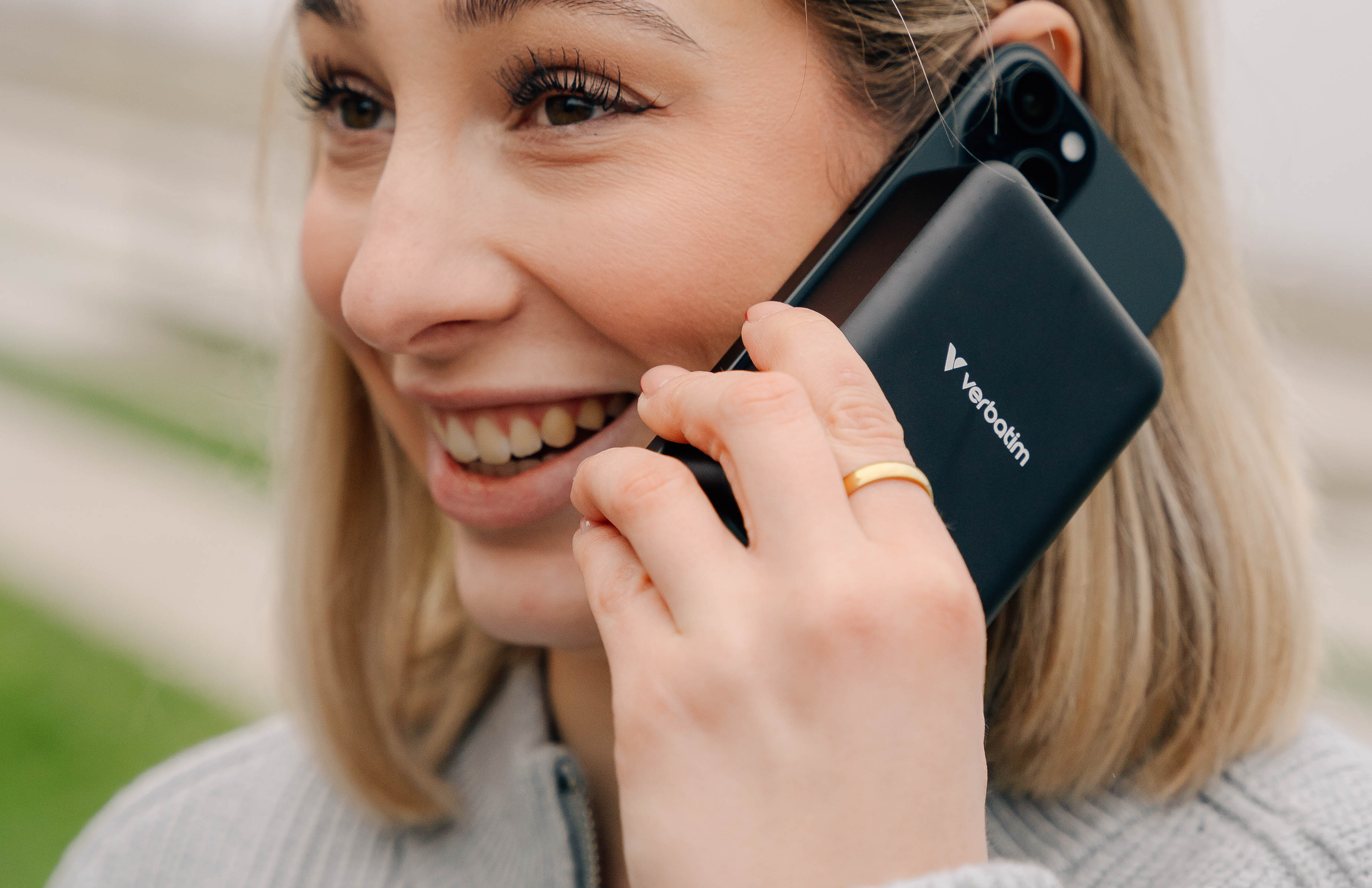 Uśmiechnięta kobieta trzymająca czarny telefon i czarny power bank. Ma na sobie pierścionek.