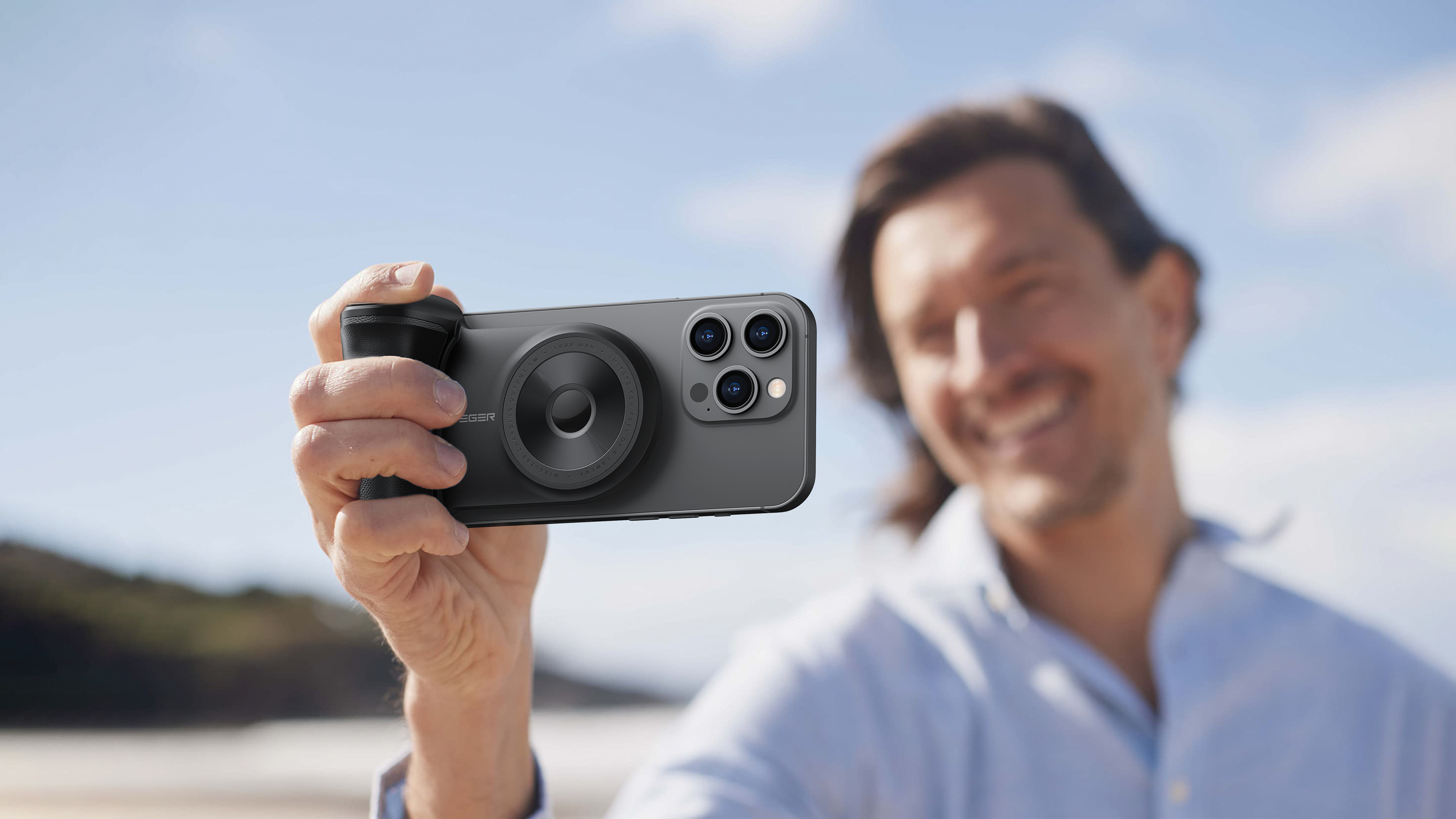 Un uomo tiene in mano un telefono con un accessorio per fotocamera, sorridendo. Lo sfondo è una spiaggia con cielo blu.