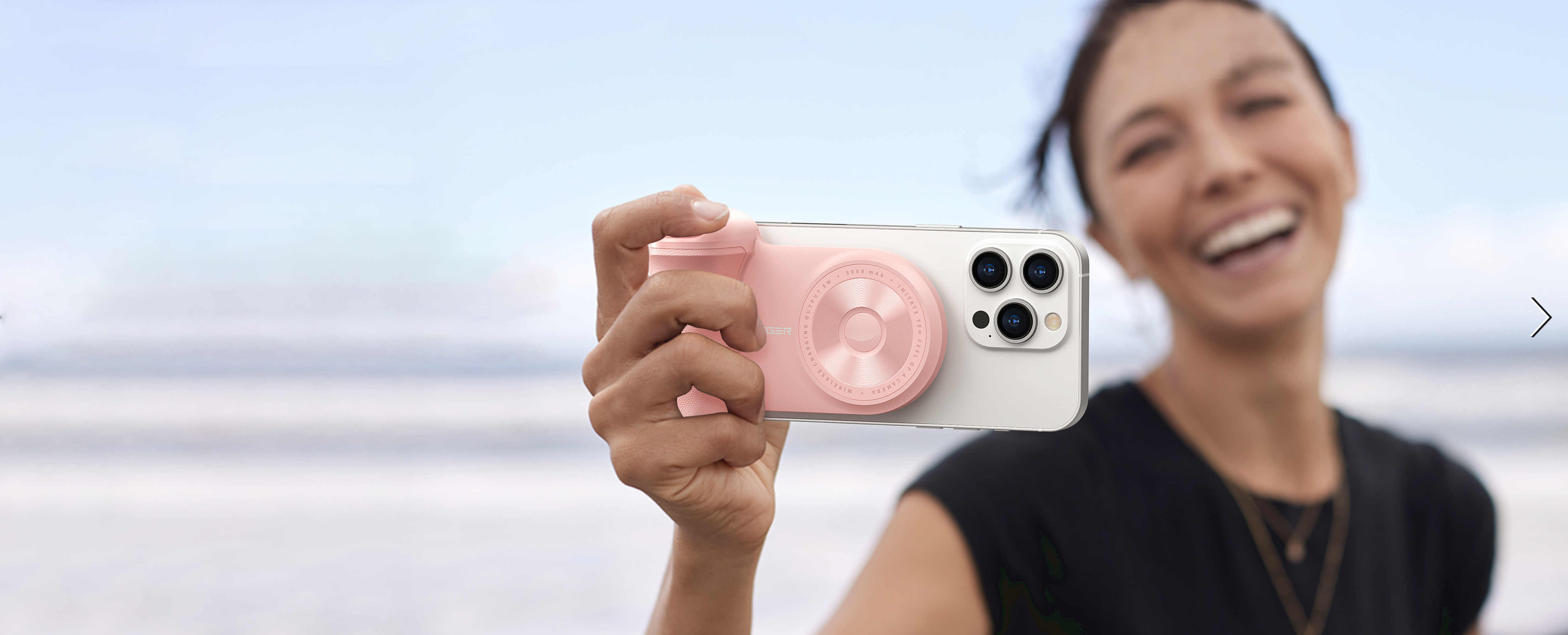 Una donna tiene un accessorio per fotocamera rosa su un telefono bianco, sorridendo alla fotocamera sulla spiaggia.