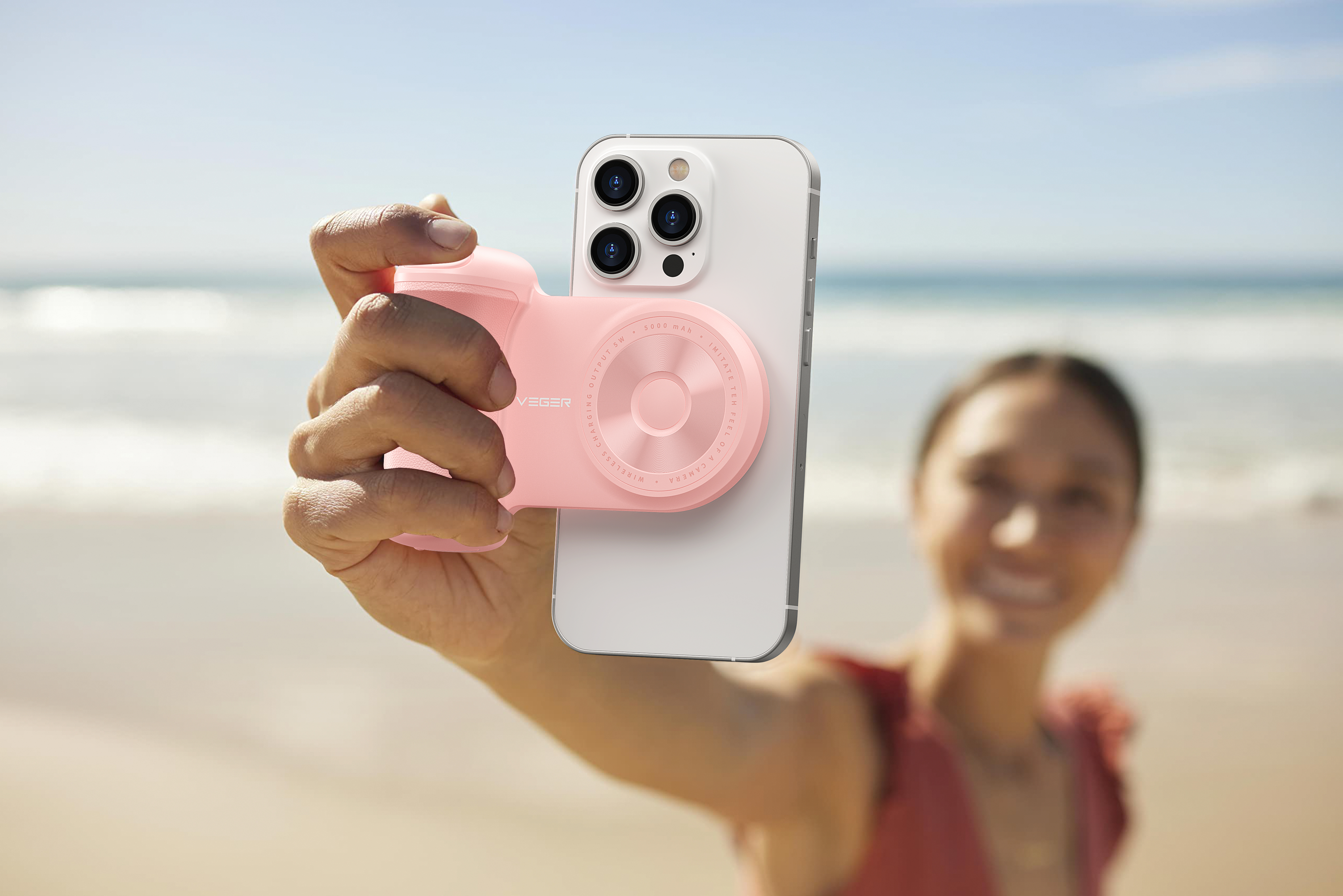 Una donna sulla spiaggia tiene un accessorio per fotocamera rosa sul telefono e sorride allo spettatore.
