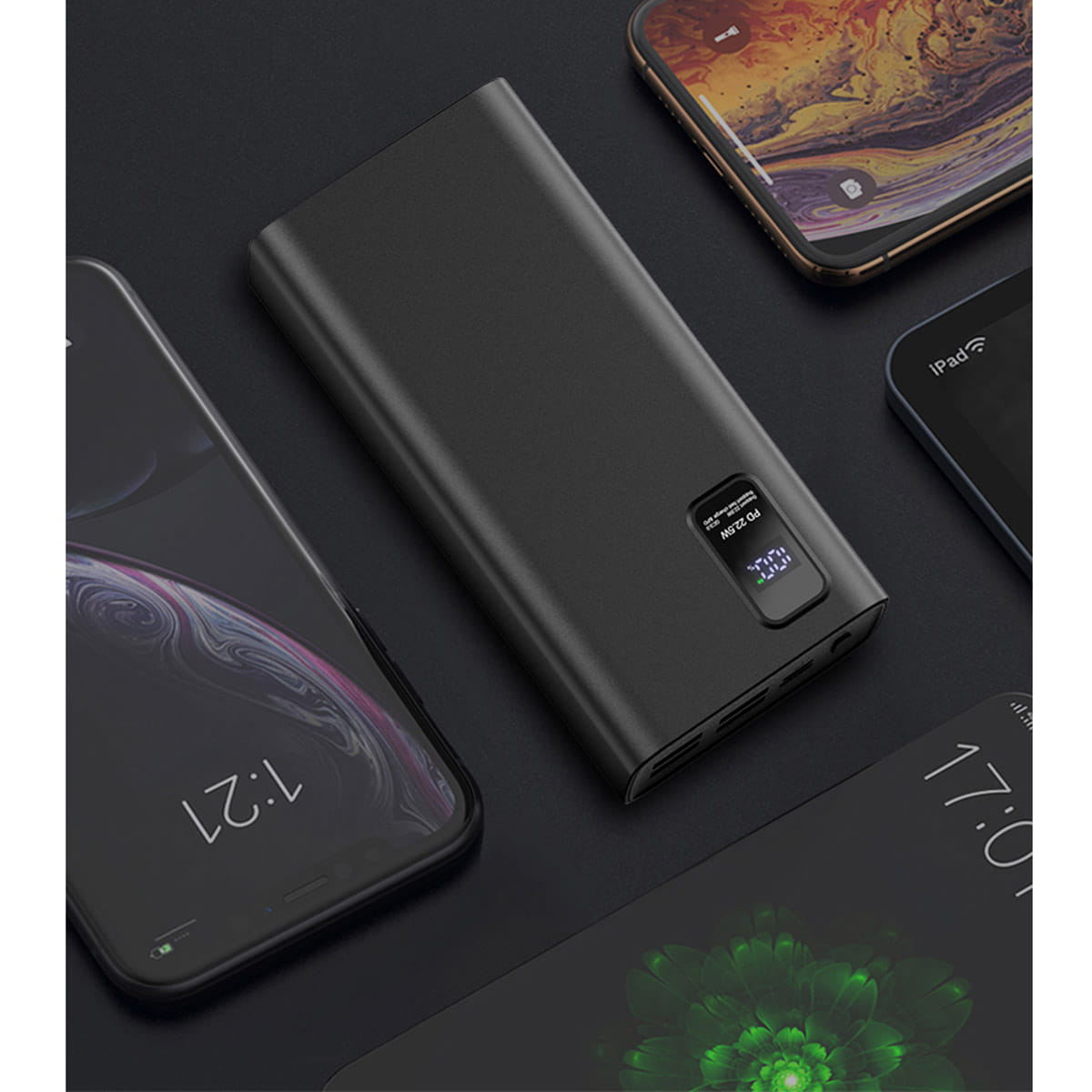 Schwarze Powerbank auf dunkler Oberfläche, umgeben von Telefonen und einem iPad. Das Display zeigt 100%.
