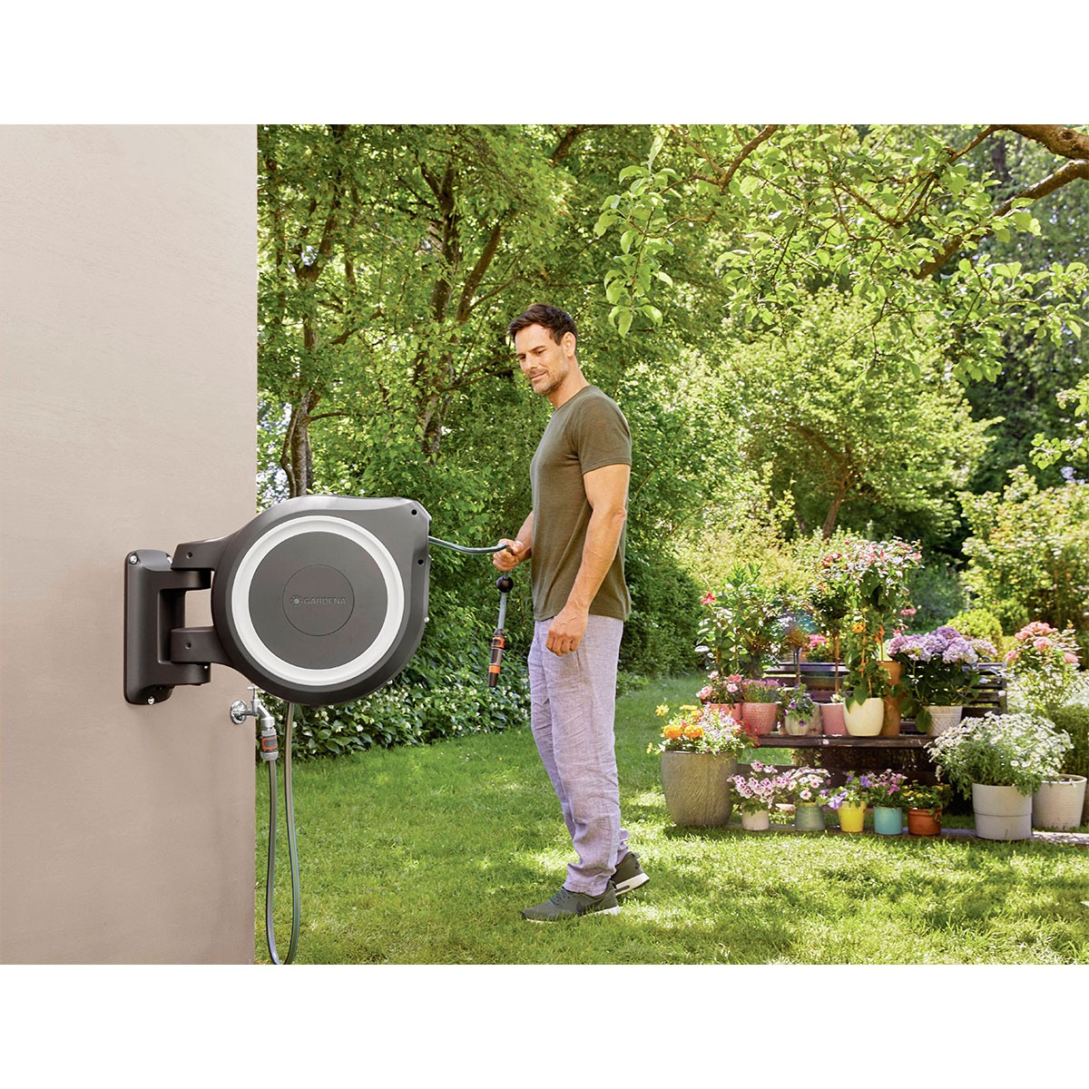 Ein Mann benutzt eine an der Wand befestigte Schlauchtrommel. Die Szene ist im Freien mit Garten im Hintergrund.