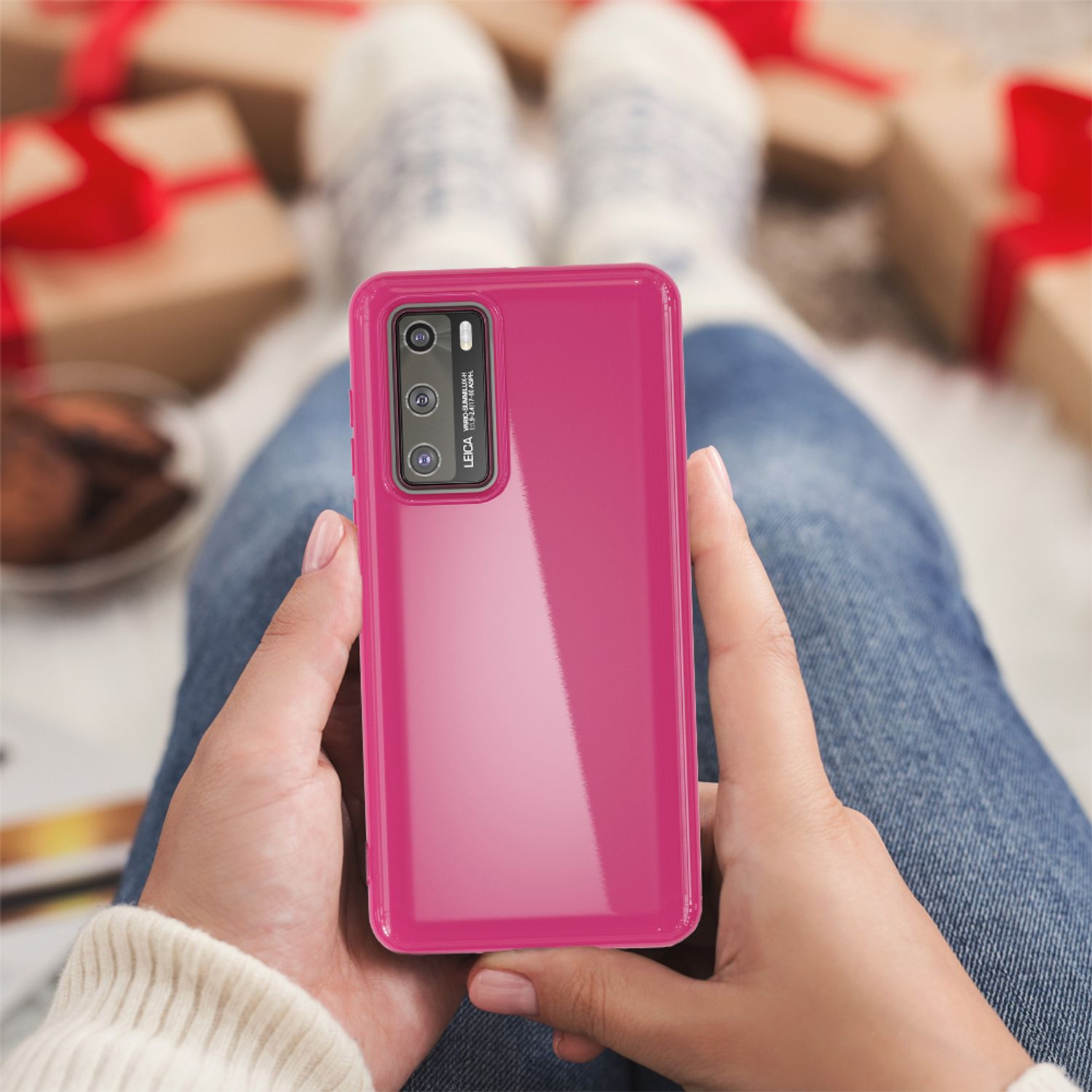 Person hält ein pinkfarbenes Telefon mit Hülle. Die Person sitzt auf einer Couch mit Geschenken und einer Decke im Hintergrund.