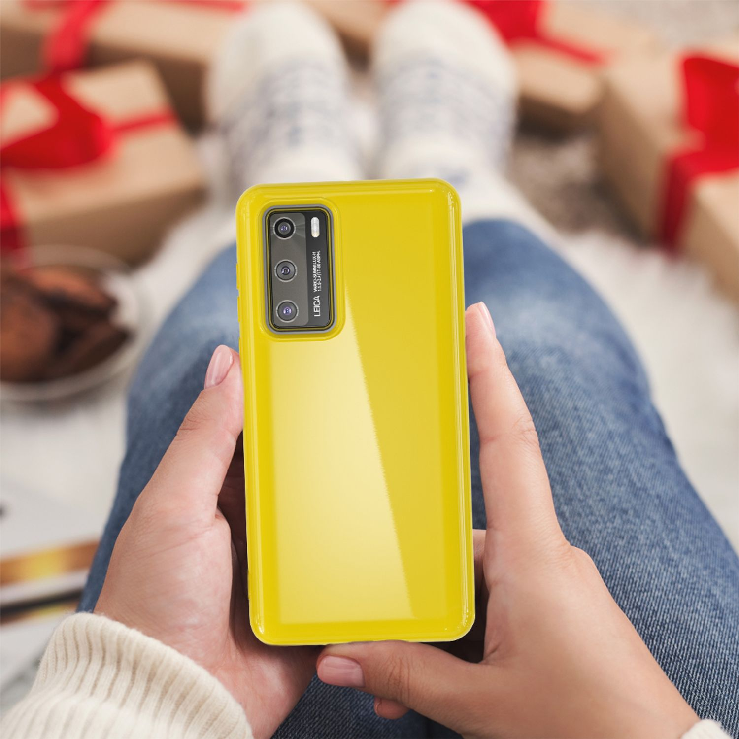 Eine Person hält eine gelbe Handyhülle. Das Telefon hat eine Kamera mit Leica-Branding. Der Hintergrund zeigt Geschenke und eine Decke.