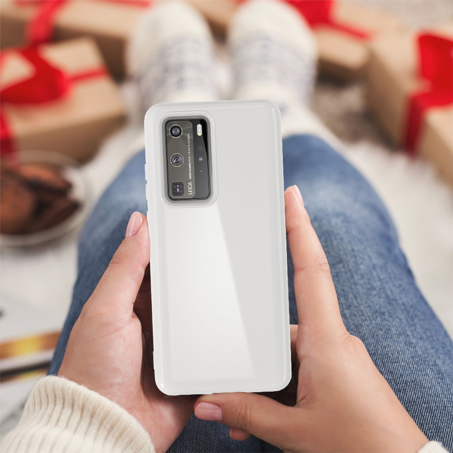 Hände halten ein Telefon in einer transparenten Hülle, sitzen auf einer Couch. Geschenke im Hintergrund.