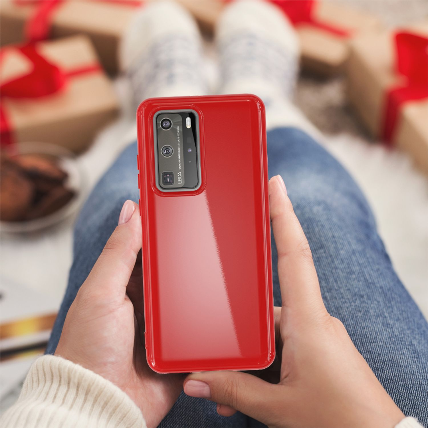 Eine Person hält ein rotes Telefon mit Kamera, trägt einen weißen Pullover, Geschenke im Hintergrund.