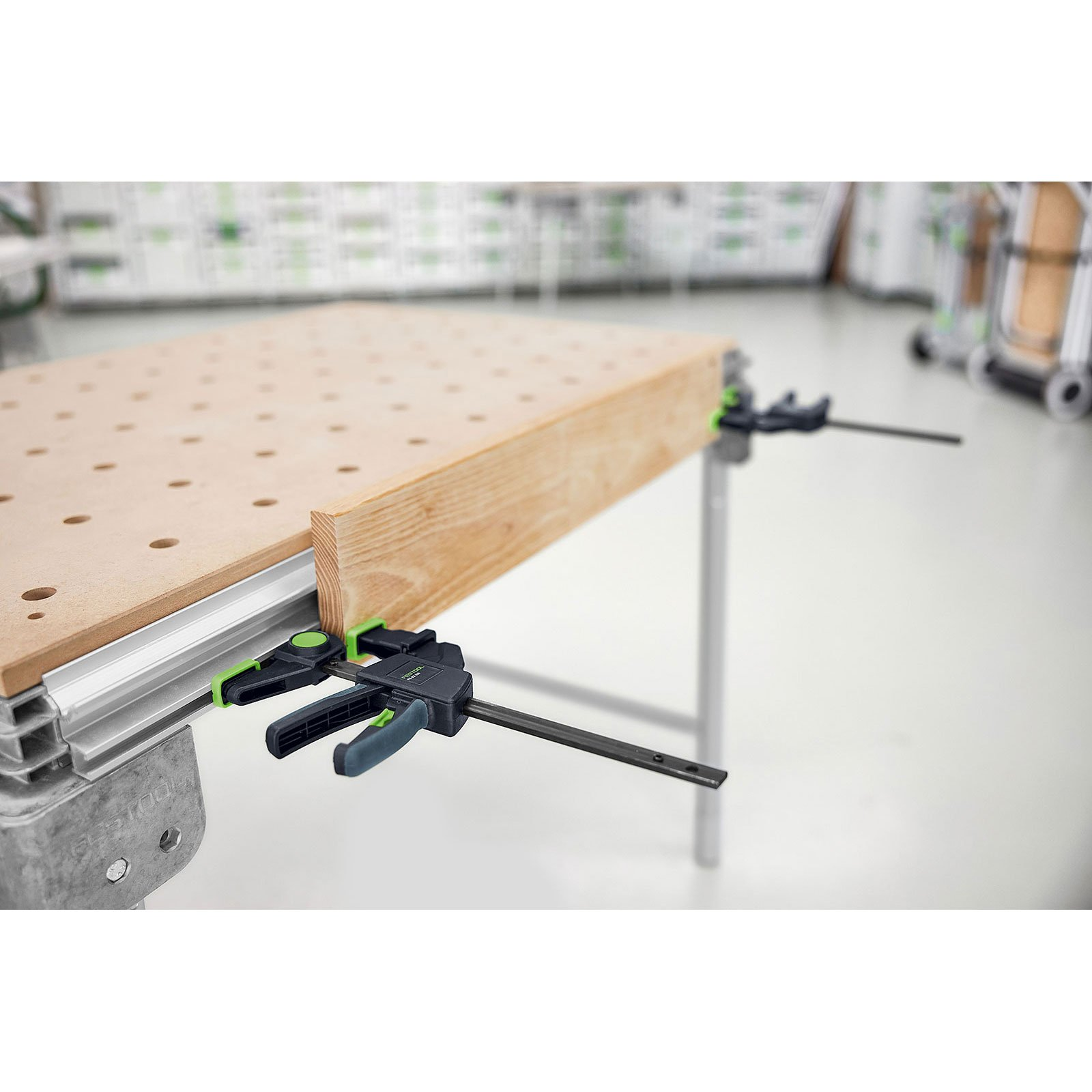 Ein Holzbrett wird mit zwei schwarz-grünen Klemmen an einer Werkbank befestigt. Die Werkbank ist silberfarben.