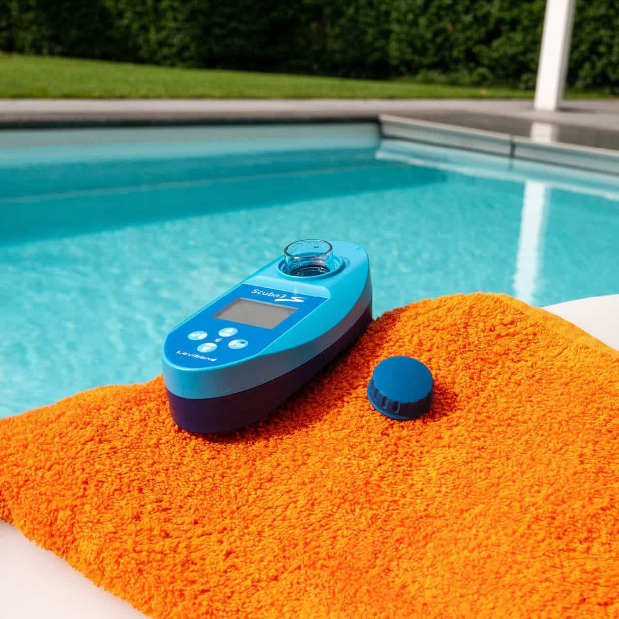 Un dispositivo azul para pruebas de agua y tapa sobre una toalla naranja junto a una piscina. El dispositivo tiene botones y una pantalla.