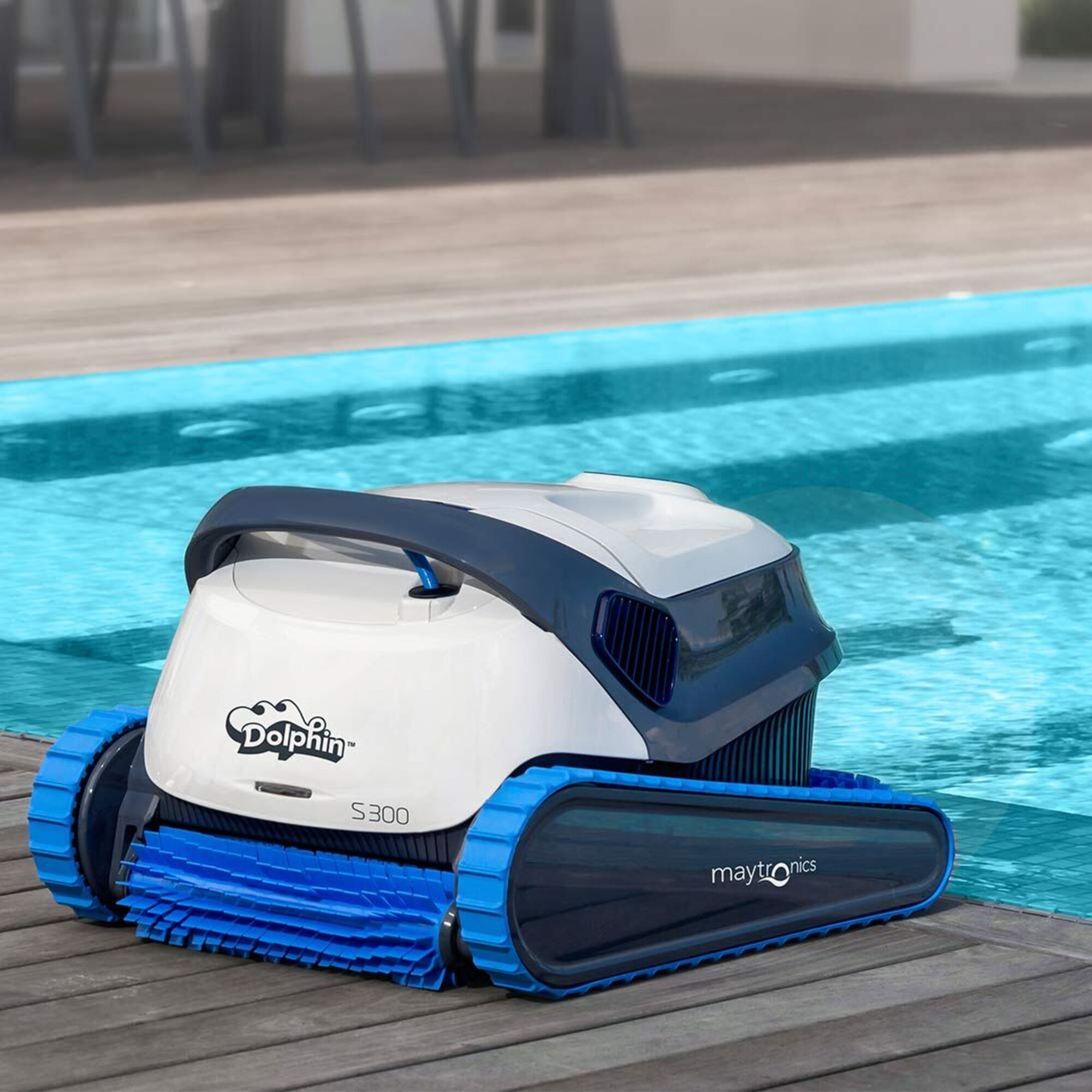 Un limpiafondos blanco y azul está sobre una cubierta de madera junto a una piscina. Tiene orugas y un cepillo azul.