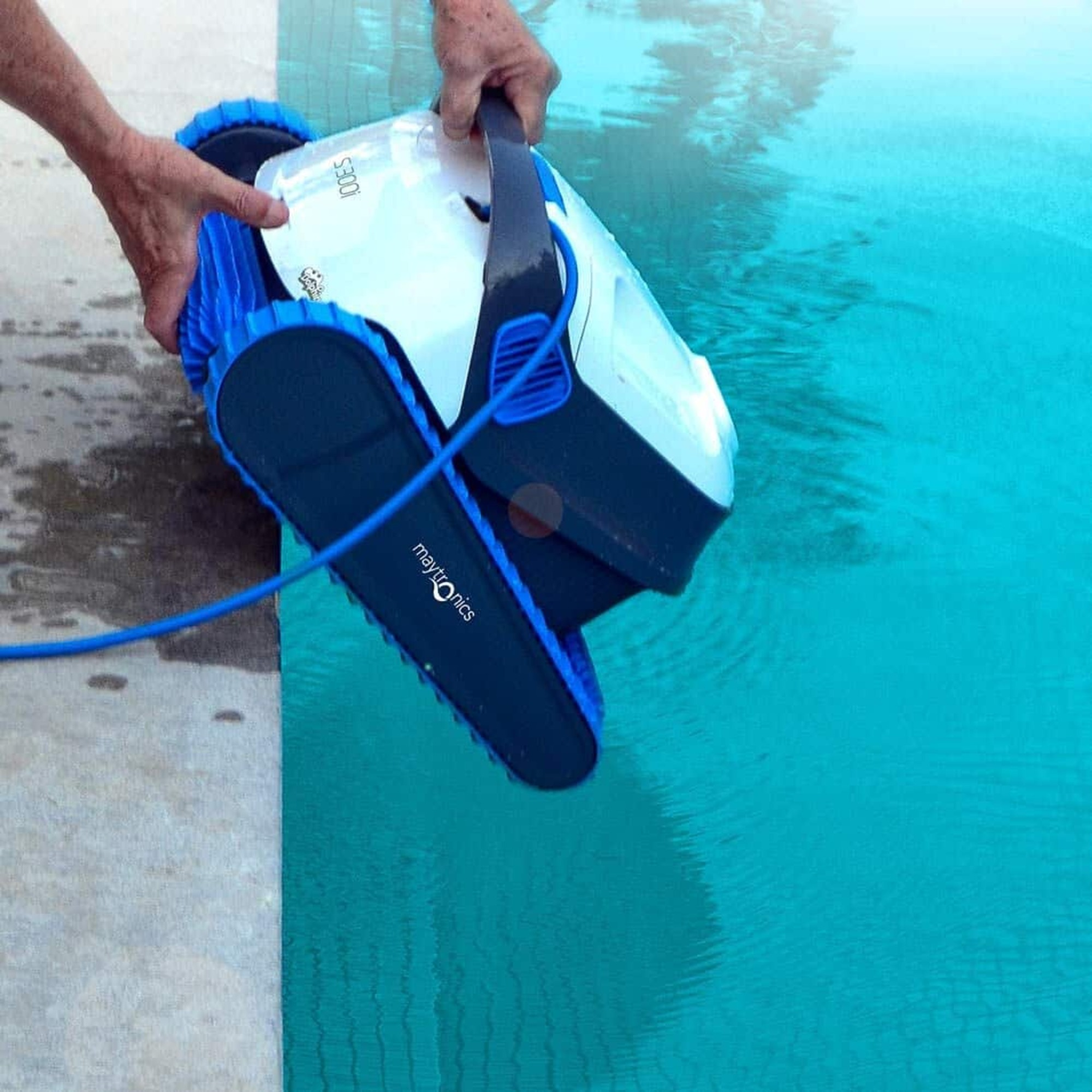 Manos sosteniendo un limpiador de piscina blanco, azul y gris sobre una piscina azul, el logo de Maytronics es visible.