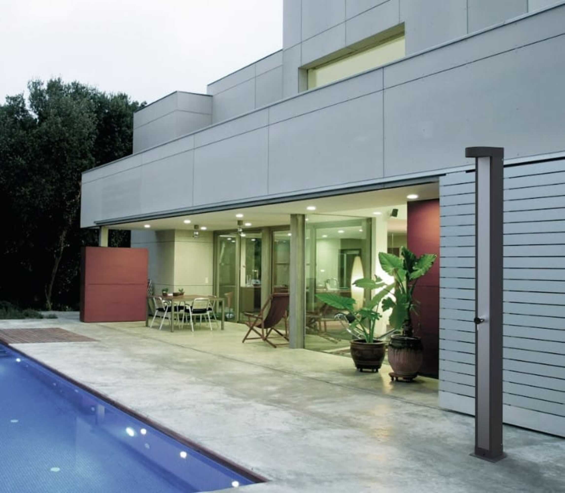 Escena al aire libre con una casa moderna, patio, piscina y una ducha exterior gris oscuro.