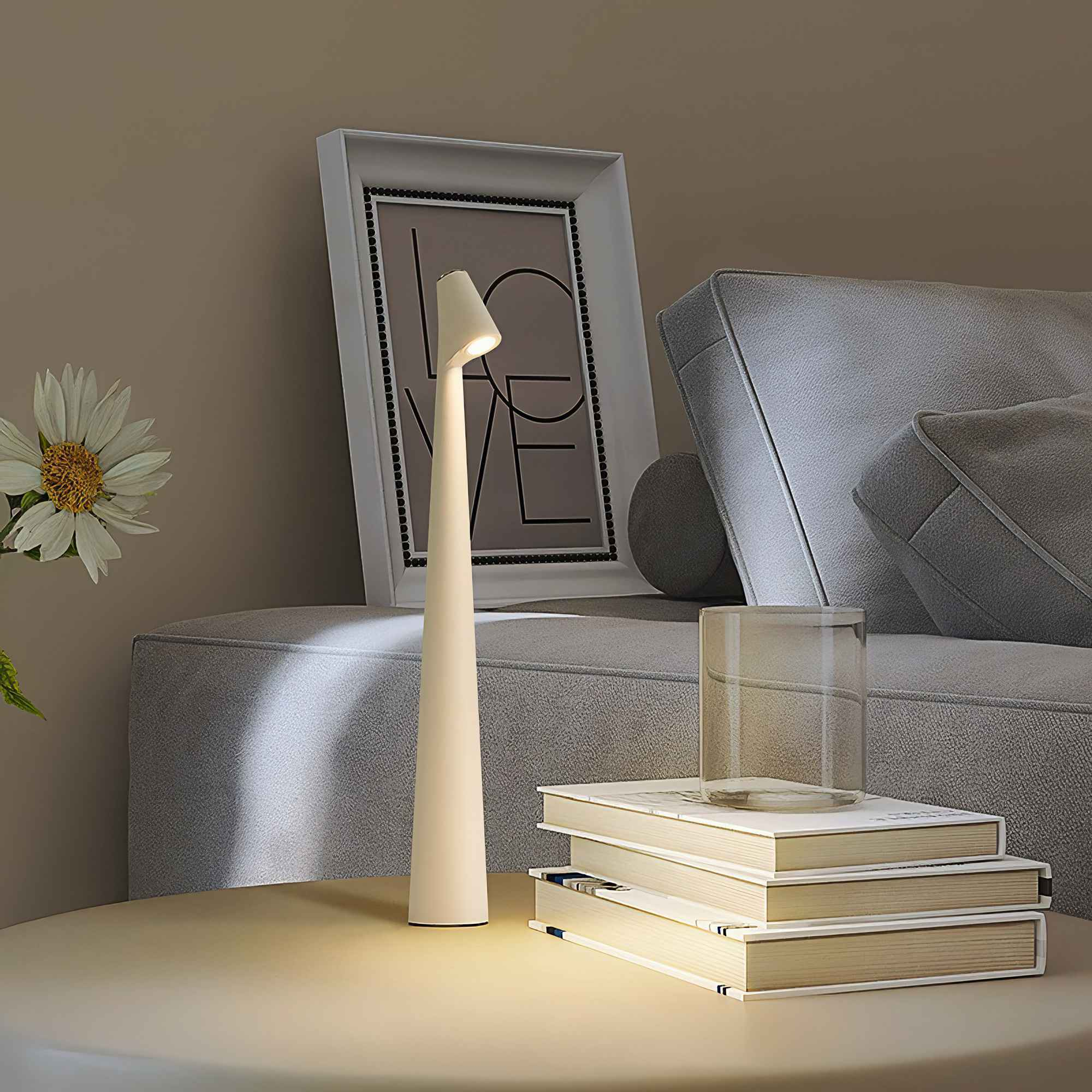 Eine moderne Lampe leuchtet auf einem Tisch neben einem Bücherstapel und einem Glas. Ein gerahmtes Bild im Hintergrund.