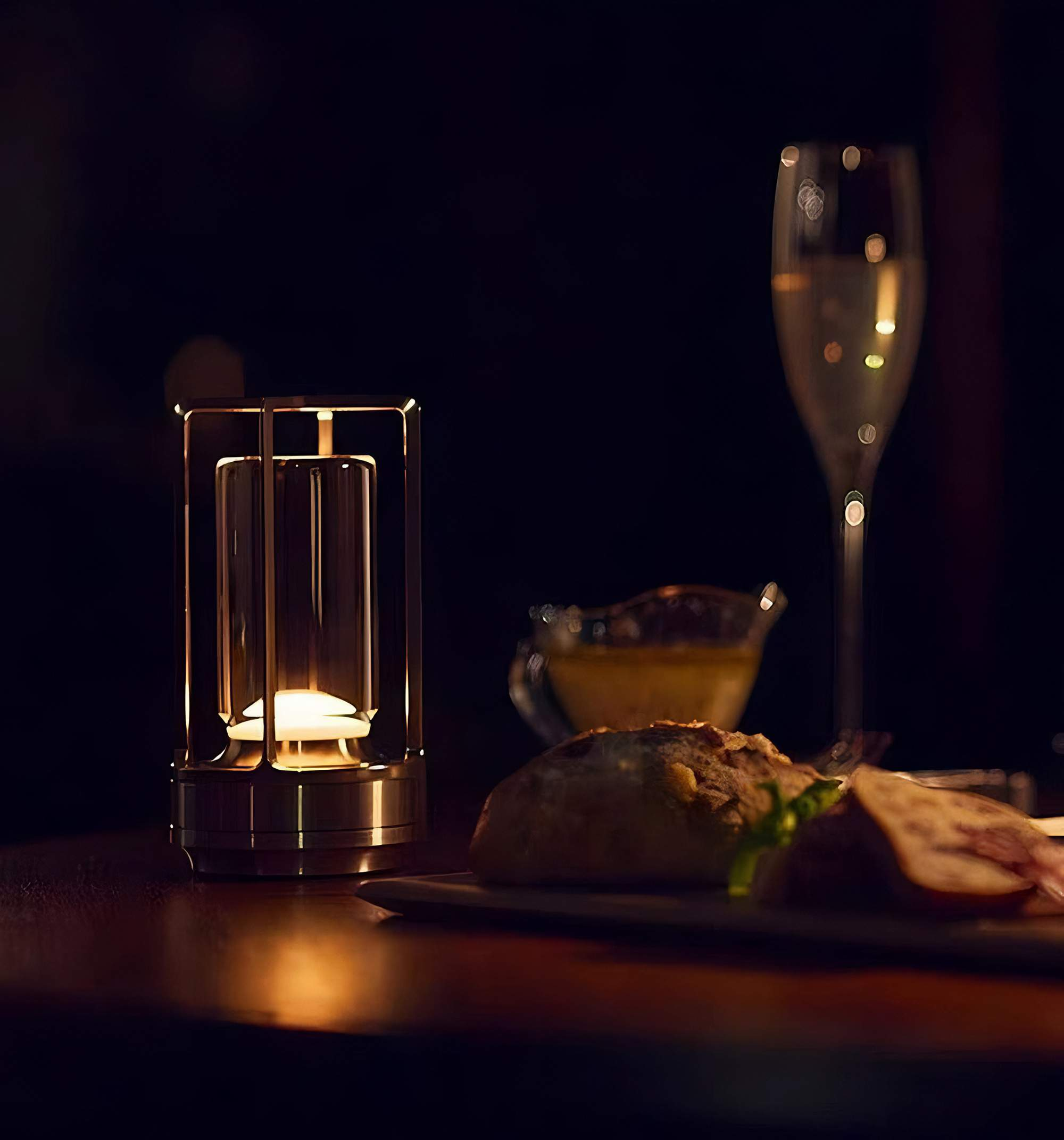 Eine beleuchtete Lampe auf einem Tisch. Im Hintergrund ein Glas Champagner, ein kleines Gericht und ein Teller mit Essen.