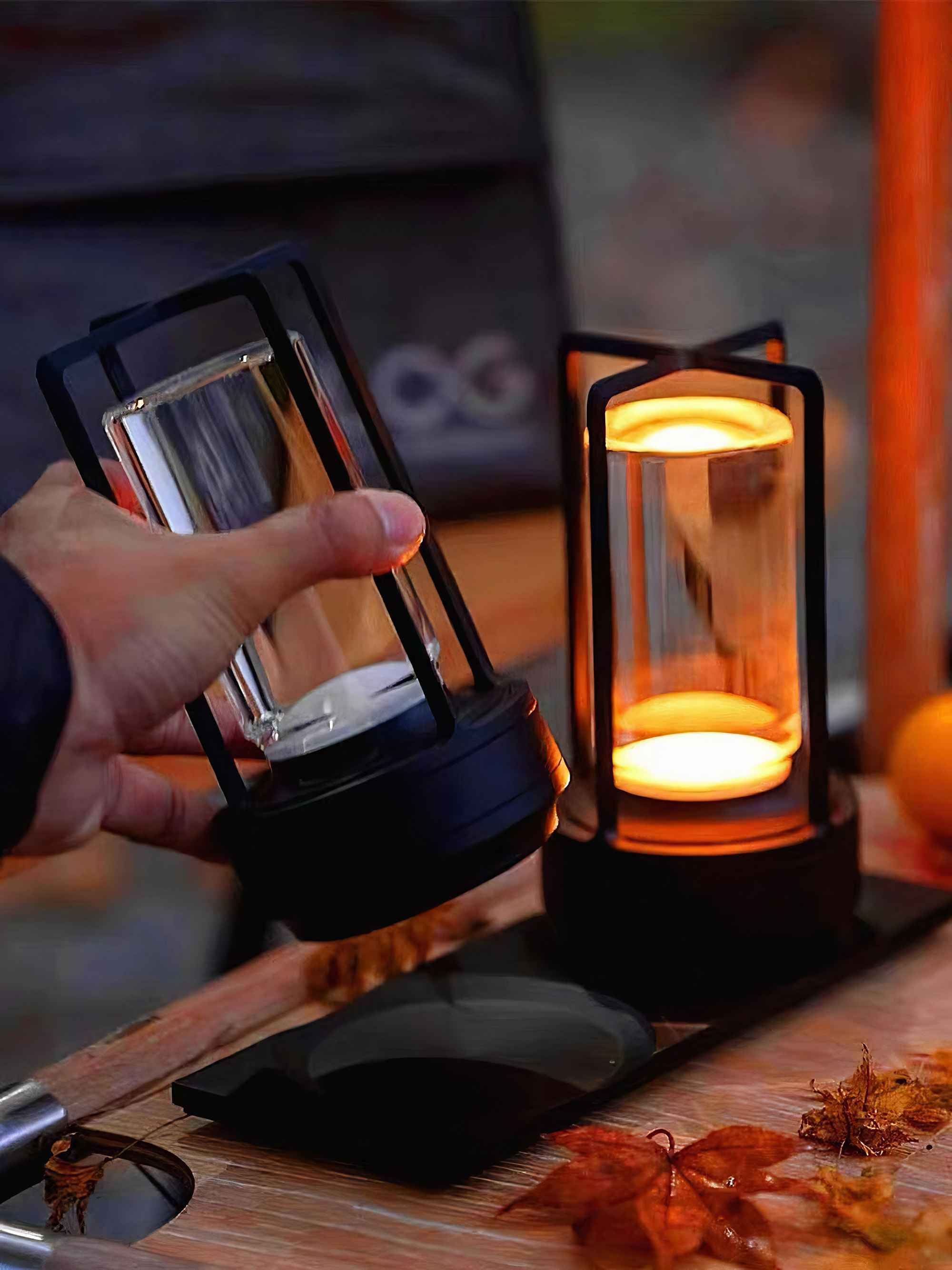 Eine Hand hält eine schwarze Laterne mit einem durchsichtigen Glaszylinder. Eine weitere beleuchtete Laterne steht auf einer Holzoberfläche.