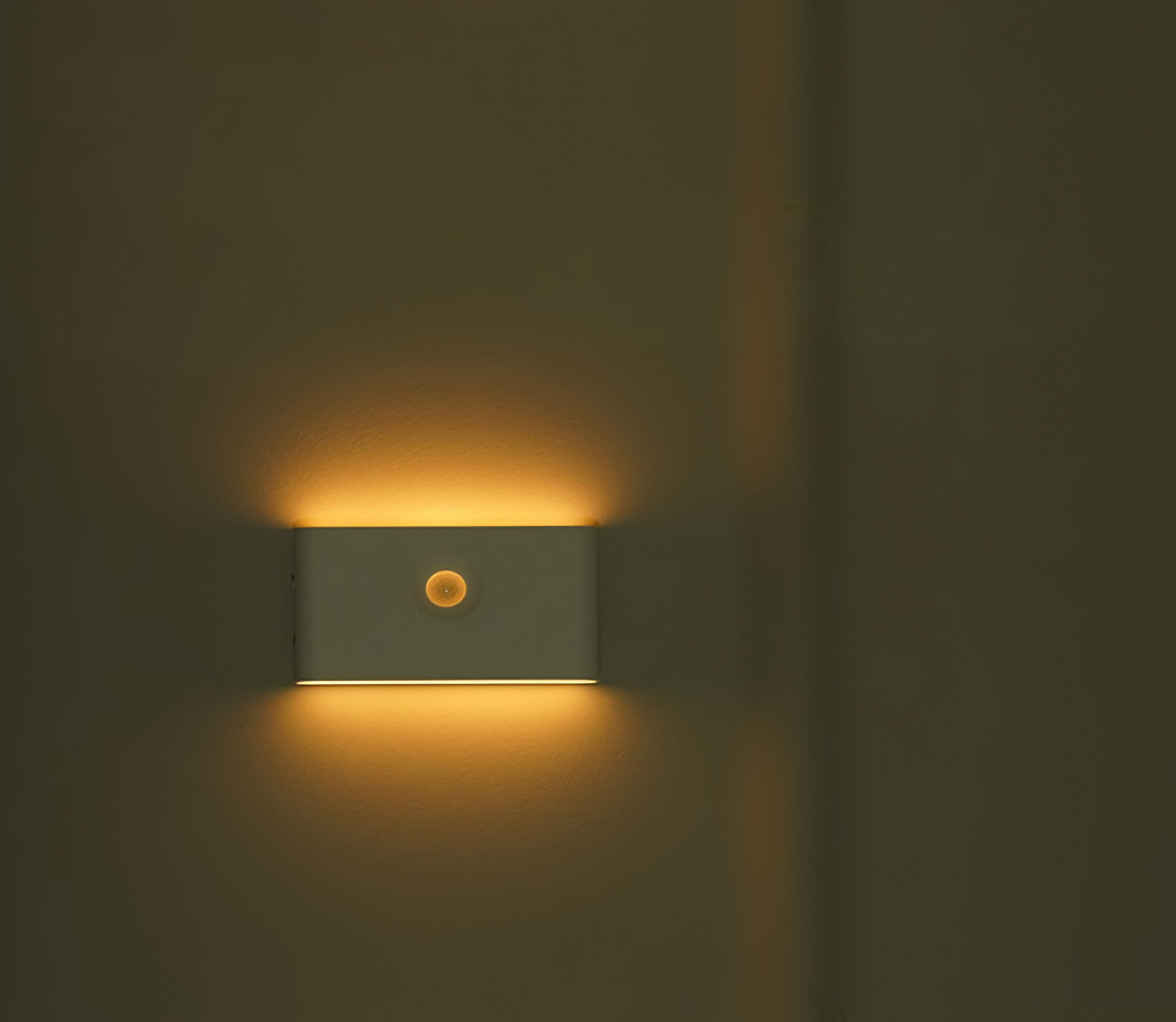 Eine rechteckige, weiße Wandleuchte, die warmes Licht ausstrahlt. Sie ist an einer beigen Wand montiert.