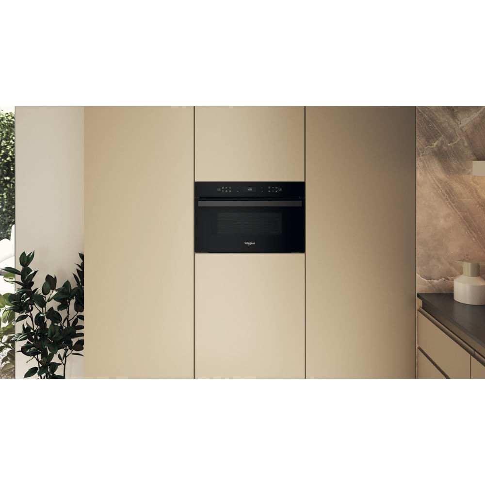 Un horno microondas negro está centrado en una pared beige. Hay armarios a ambos lados. Una planta a la izquierda.