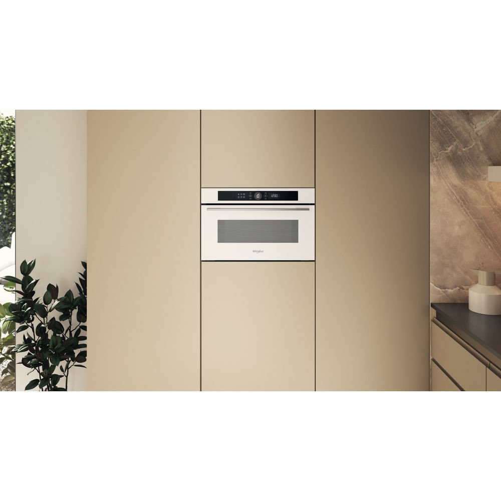 Un forno a microonde bianco è integrato in un armadio beige. Una pianta a sinistra, una finestra a sinistra.
