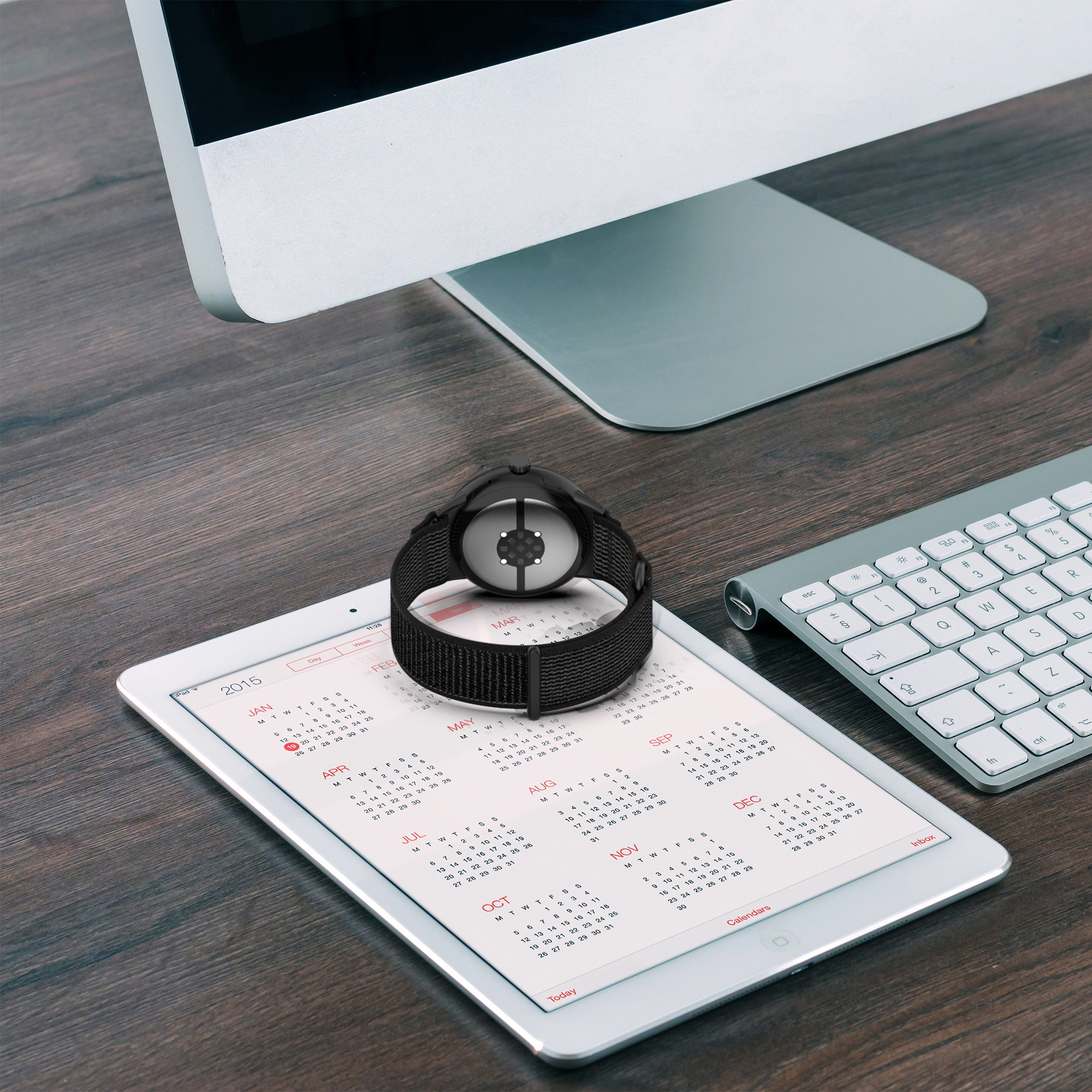 Eine schwarze Uhr auf einem Tablet mit Kalender, im Hintergrund eine Tastatur und ein Monitor.