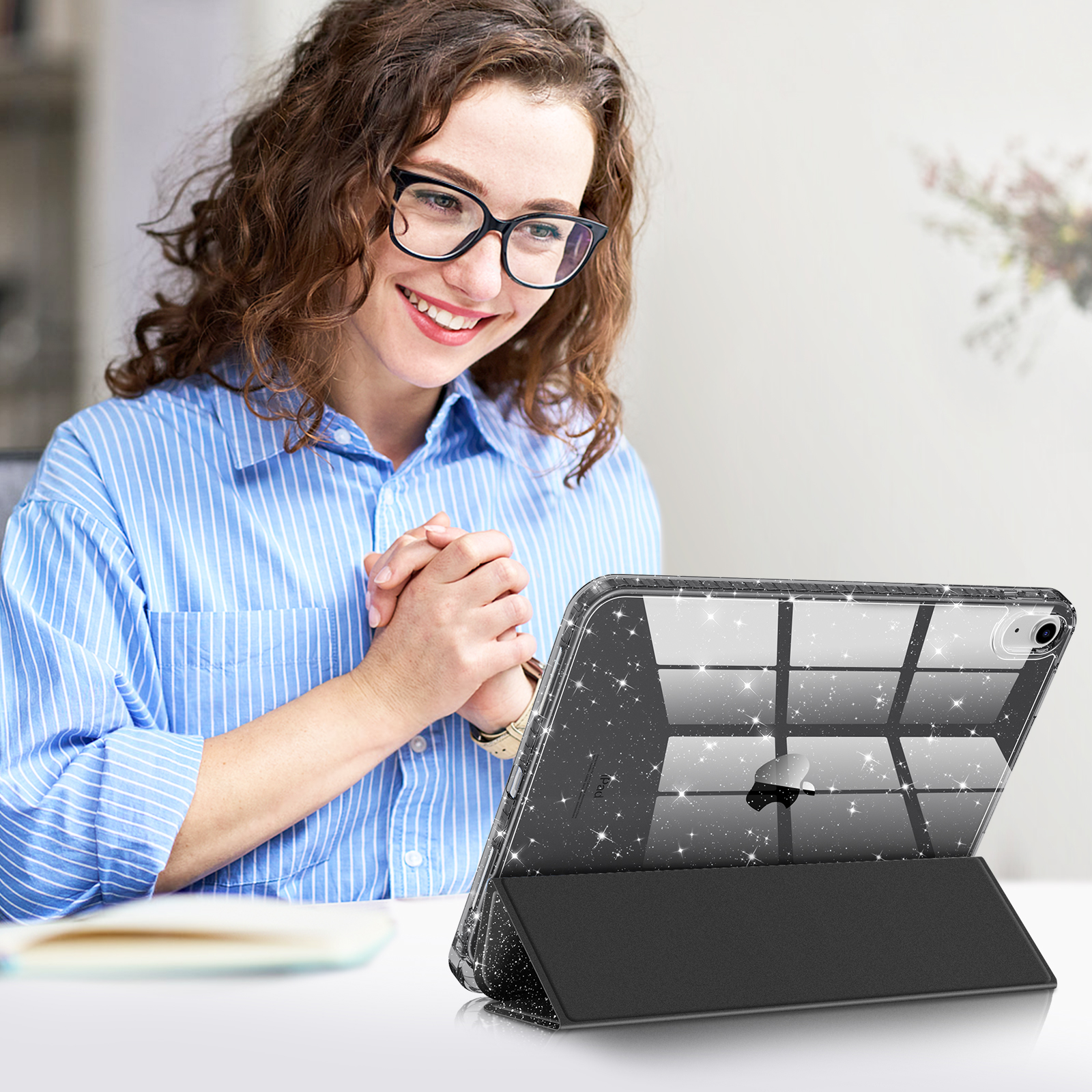Eine Frau lächelt und schaut auf ein iPad. Das iPad befindet sich in einer schwarzen Hülle. Sie sitzt drinnen an einem Tisch.