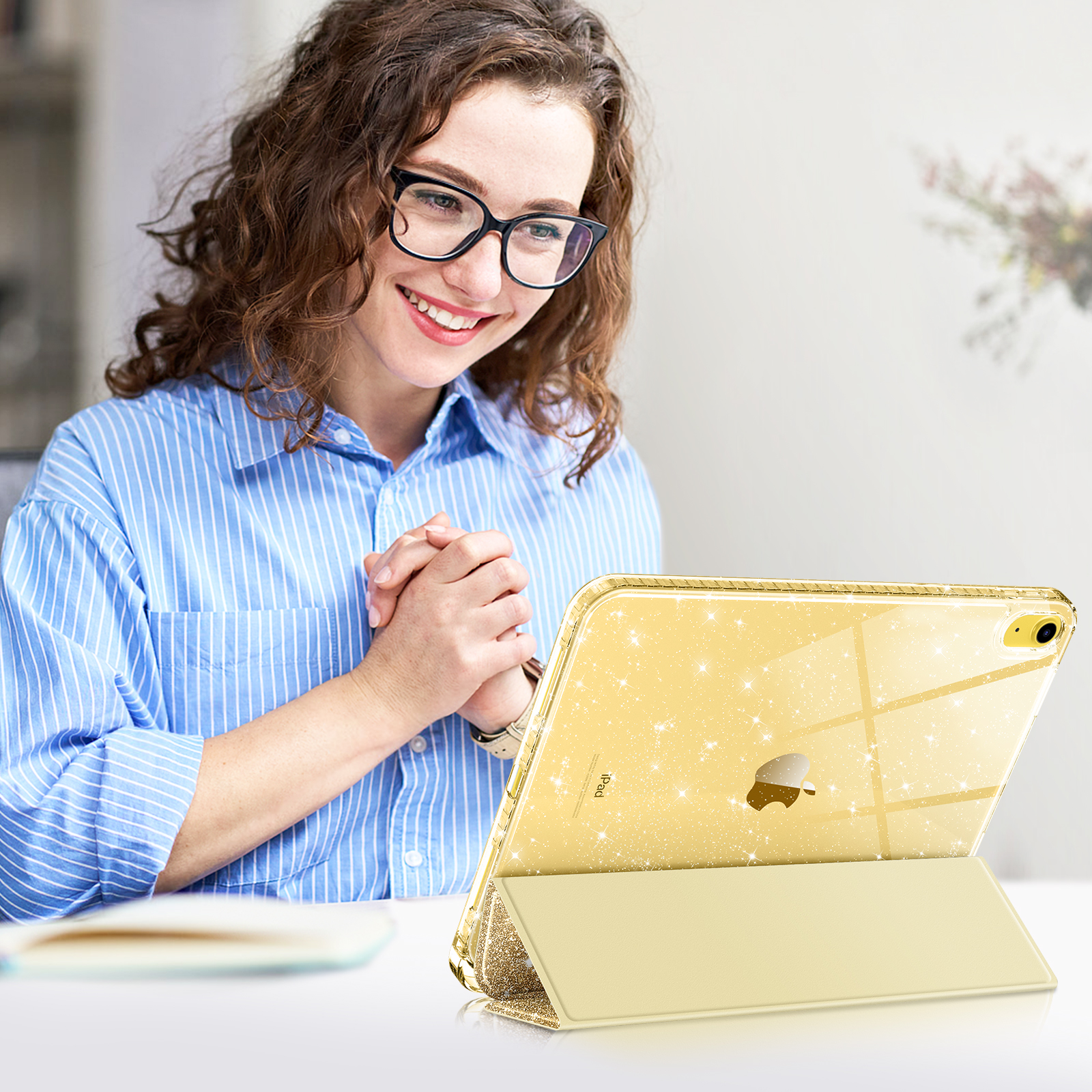 Frau mit Brille hält ein iPad in einer goldenen, glitzernden Hülle. Das iPad steht im Ständermodus auf einem Tisch.