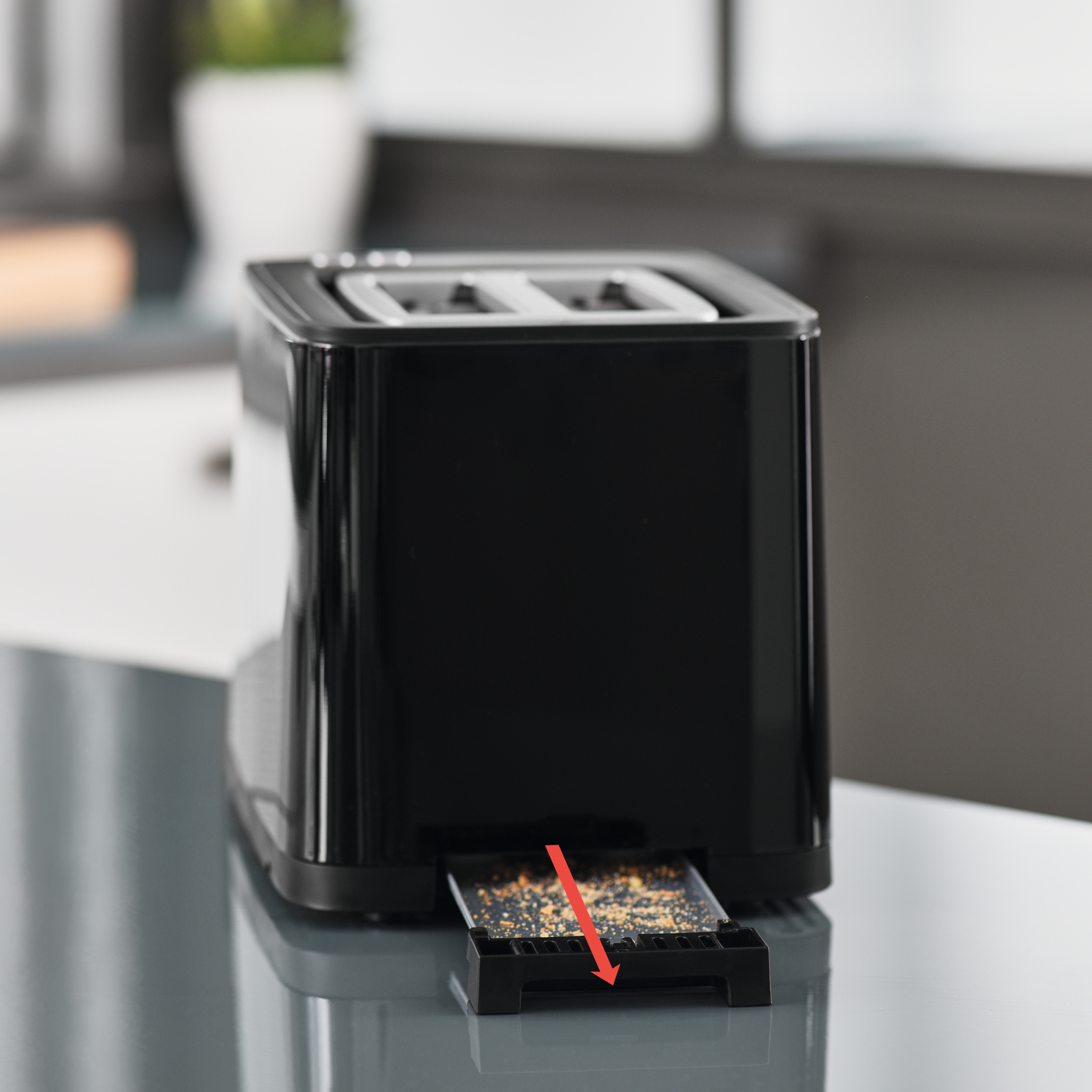 Czarny toster z wysuniętą tacą na okruchy. Taca jest pełna okruchów. Czerwona strzałka.