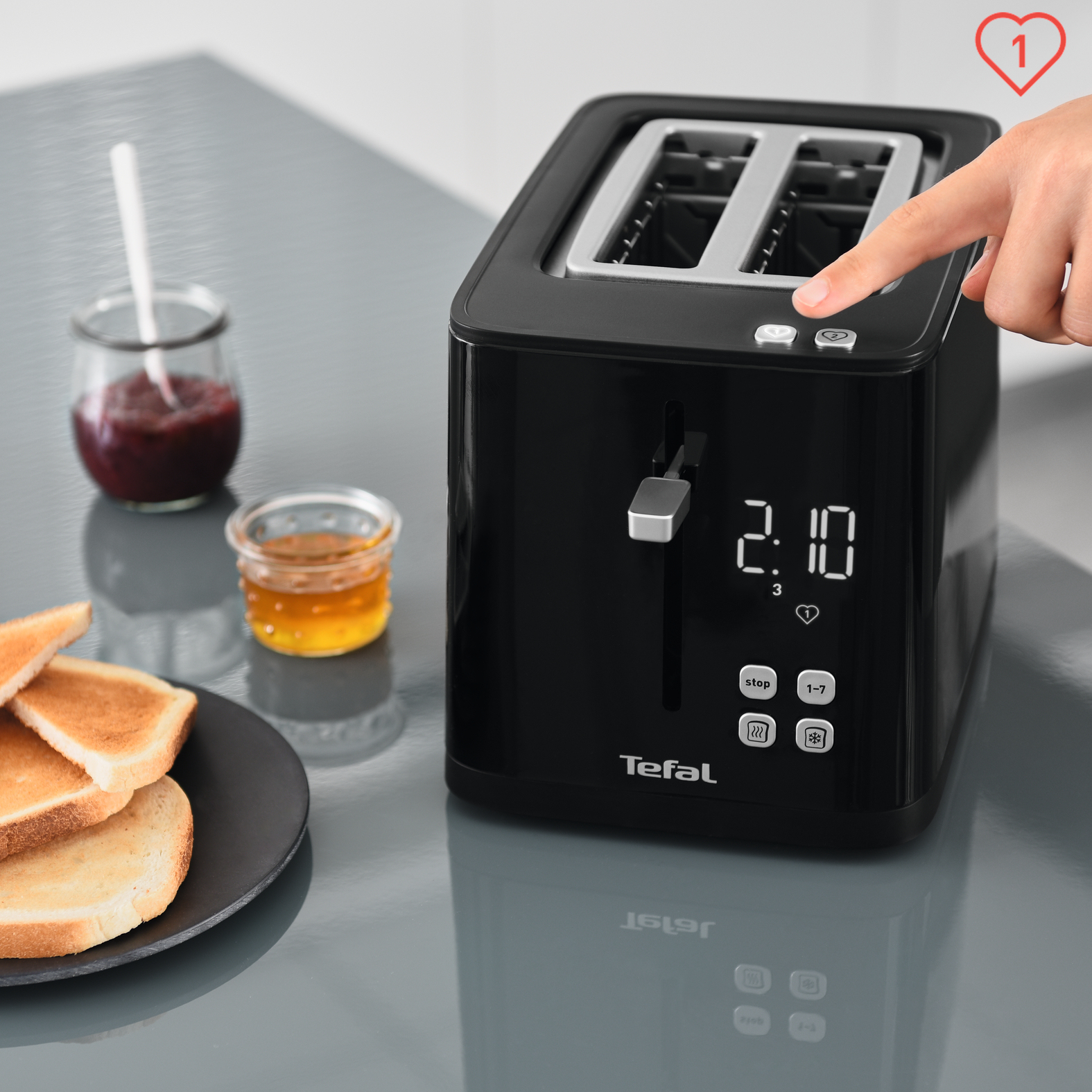 Czarny toster Tefal z tostami, dżemem i miodem. Ręka naciska przycisk z ikoną serca.