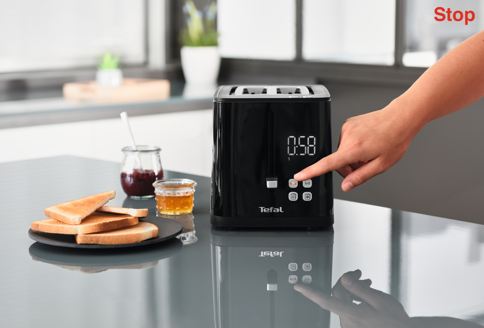 Ręka naciska przycisk na czarnym cyfrowym tosterze obok tostów, dżemu i miodu na stole w kuchni.