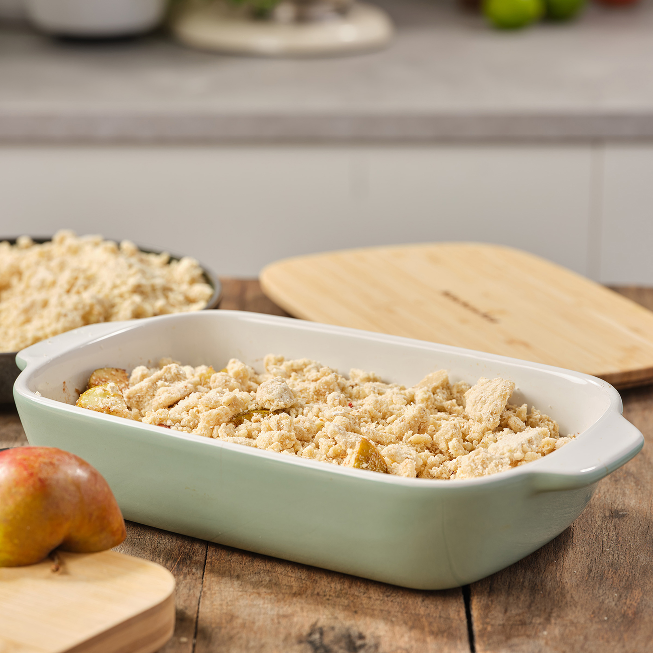 Backform mit Streuseltopping, Apfelscheiben und einer Birne. Ein Holzbrett und ein KitchenAid-Deckel sind im Hintergrund.