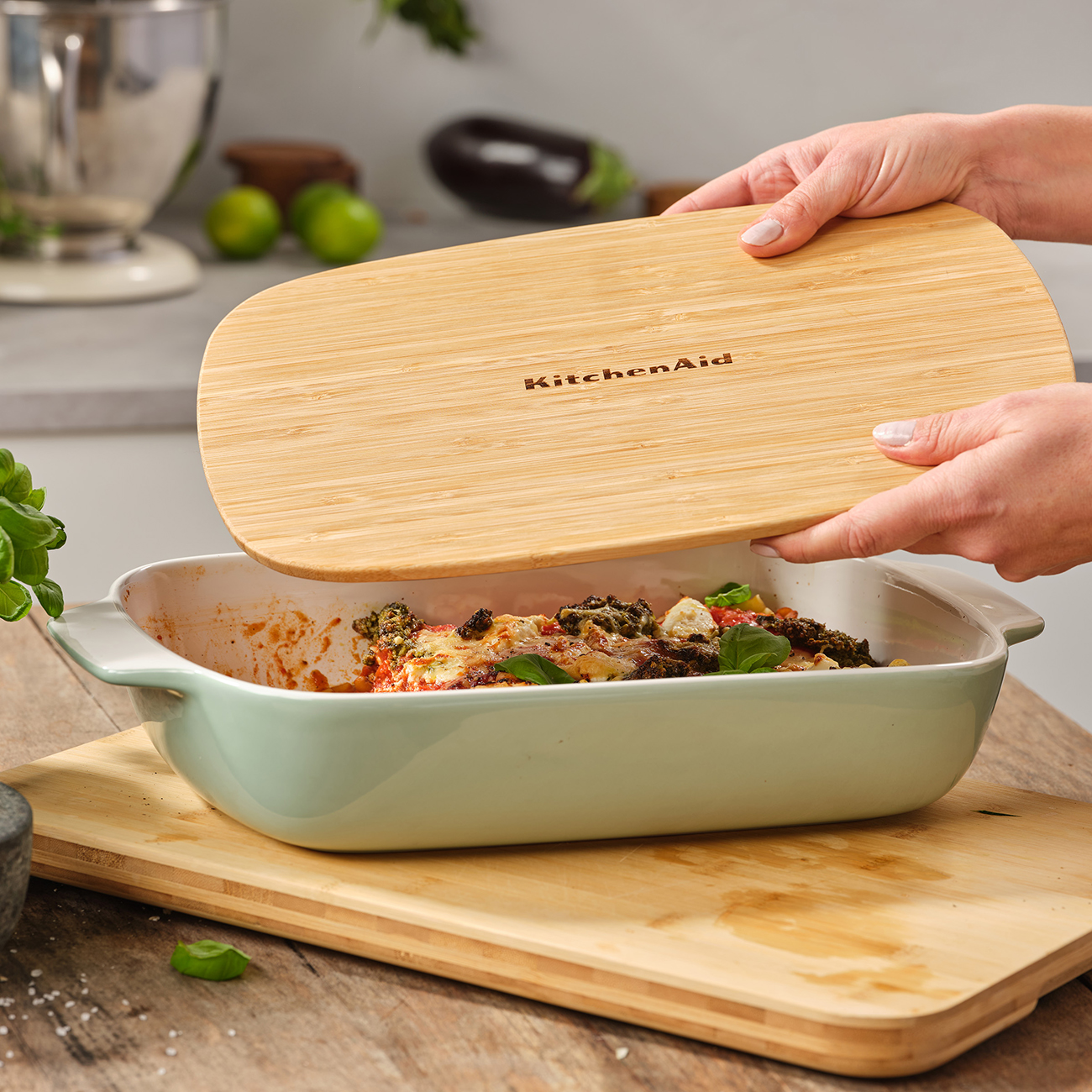 Person öffnet einen Deckel einer Backform mit Essen. Holztisch, Küche im Hintergrund.