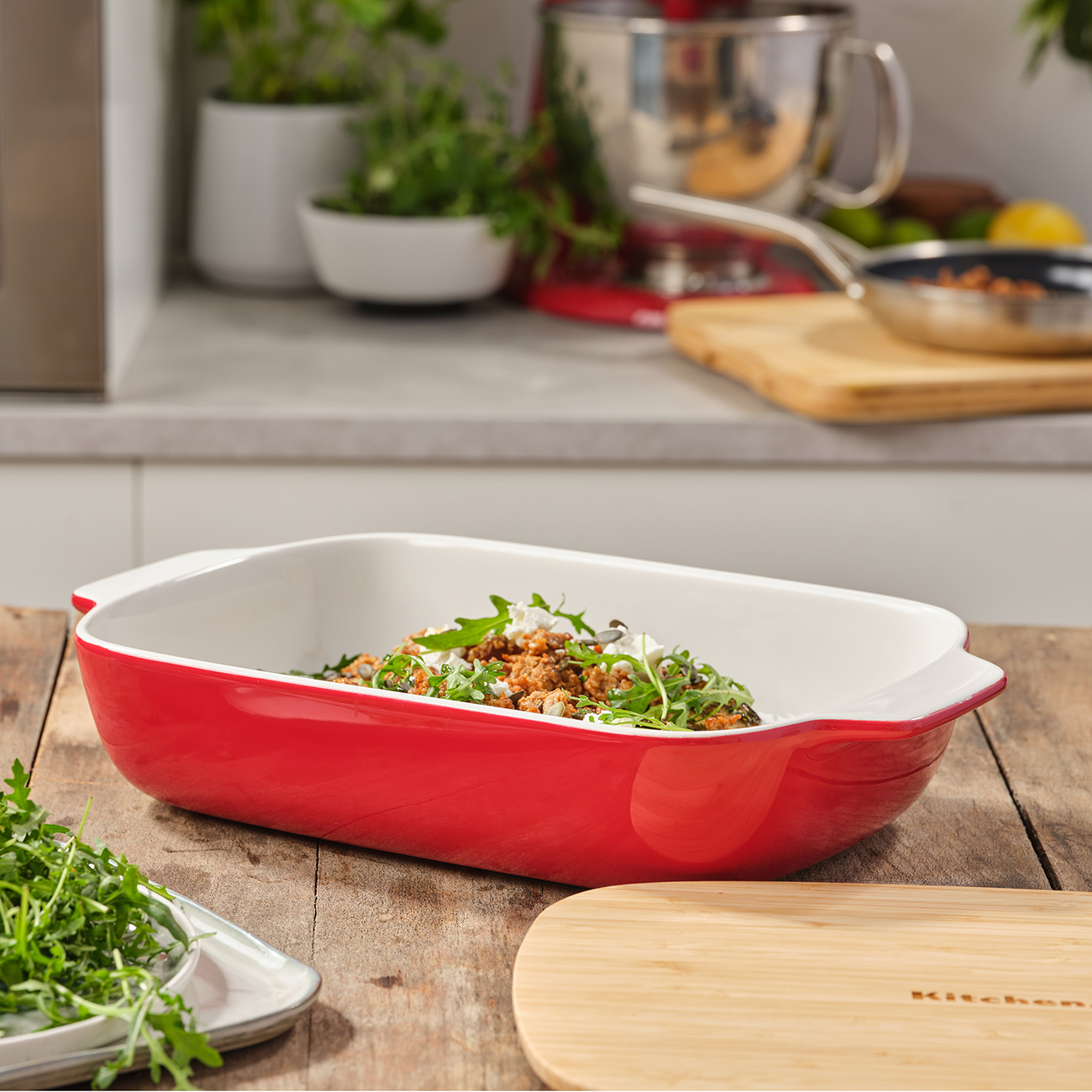 Eine rote Backform mit einer gekochten Mahlzeit. Enthält Rucola und andere Zutaten. Holztisch vorhanden.