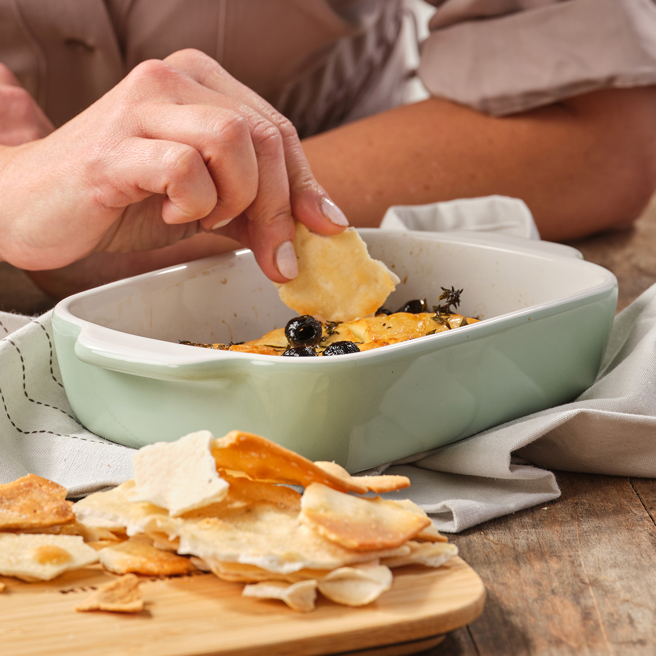 Eine Person, die einen Cracker zu einem Gericht mit gebackenem Essen und Oliven auf einem Holztisch hinzufügt.