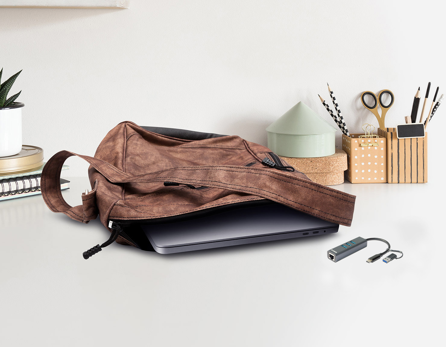 Ein brauner Rucksack ist auf einem Tisch geöffnet. Ein Laptop ist darin. Ein Hub und Kabel liegen daneben.