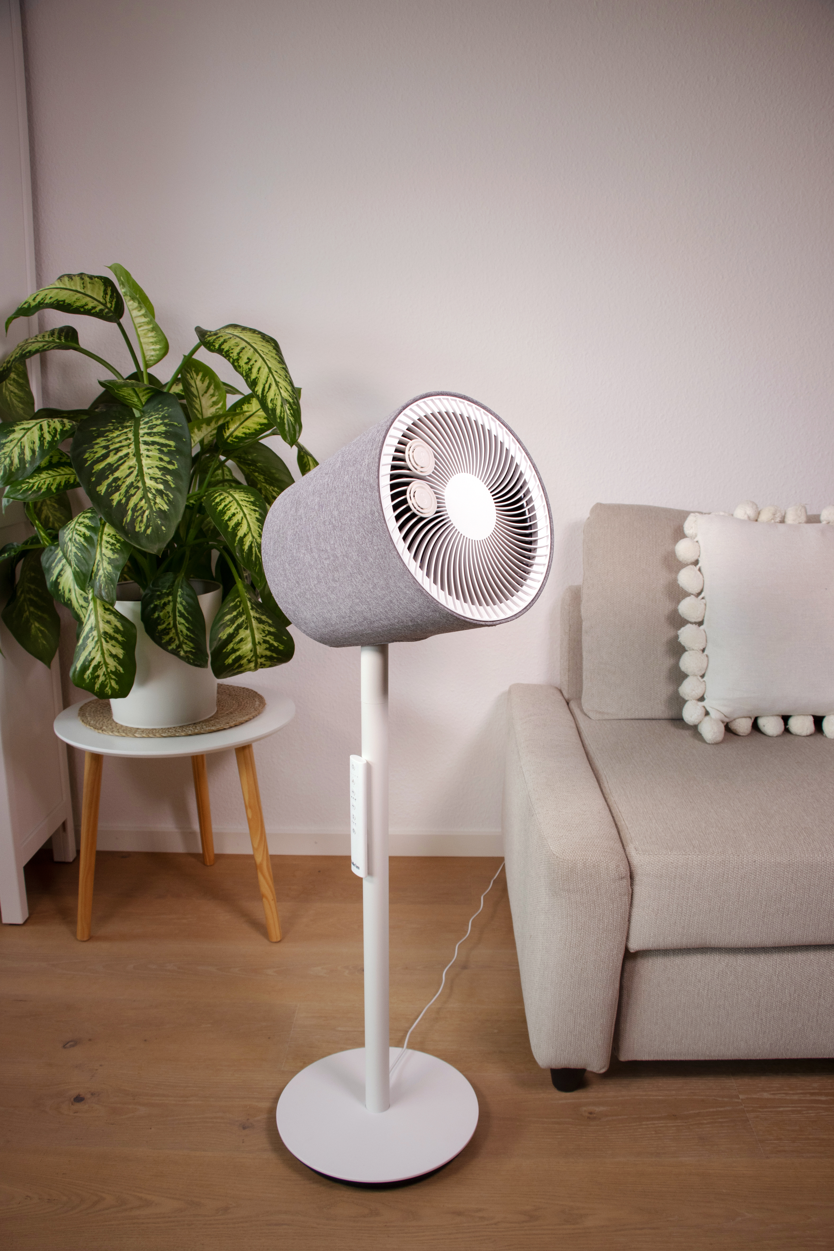 Ventilateur recouvert de tissu gris sur pied blanc. Intérieur. Base et colonne blanches. Canapé au fond.