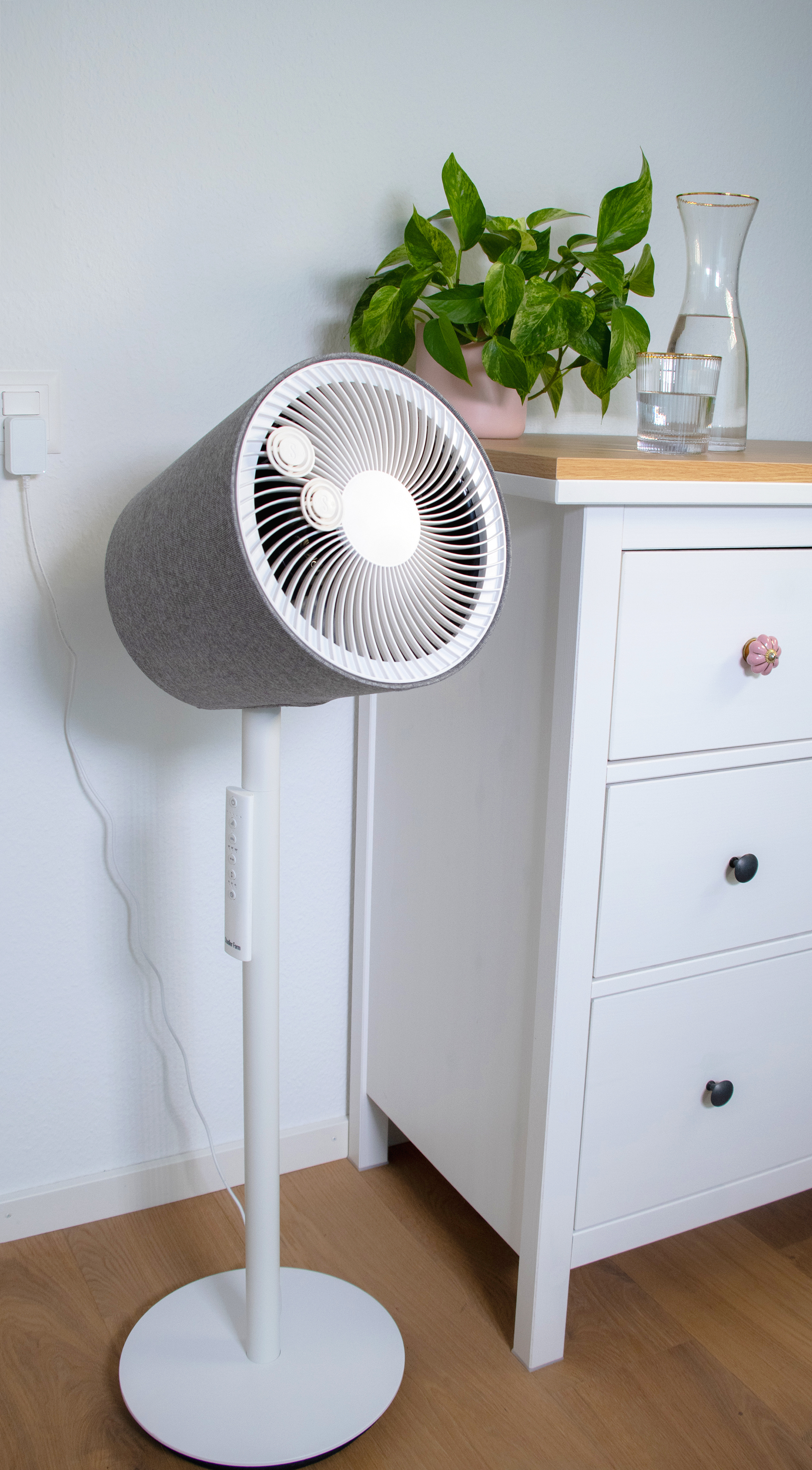 Ventilateur recouvert de tissu gris sur pied blanc. Mur blanc, commode et plante en arrière-plan.
