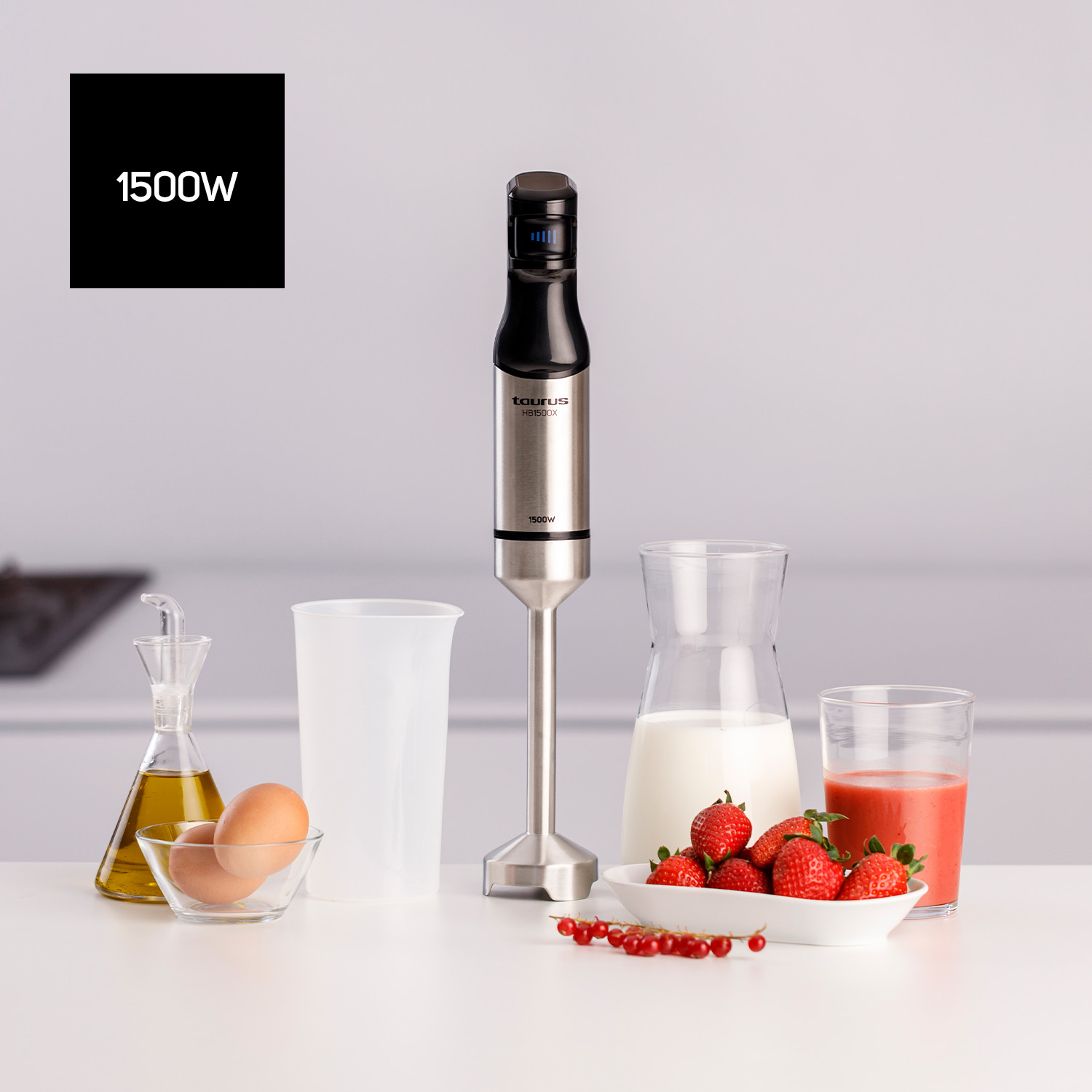 Batidora de inmersión con ingredientes: huevos, aceite, fresas, leche y grosellas rojas sobre una mesa.