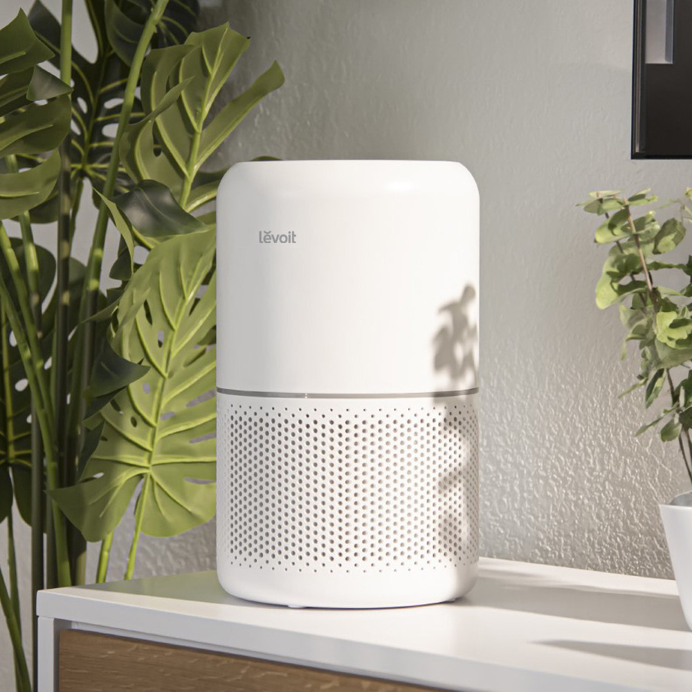 Biały oczyszczacz powietrza na białej powierzchni, z zielonymi roślinami po bokach.