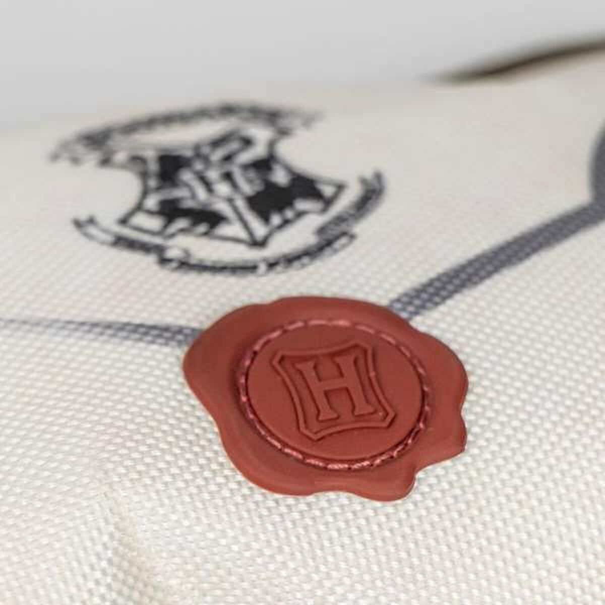 Nahaufnahme eines roten Siegels auf einem weißen Kissen mit Hogwarts-Wappen. Das Siegel hat ein 'H'.