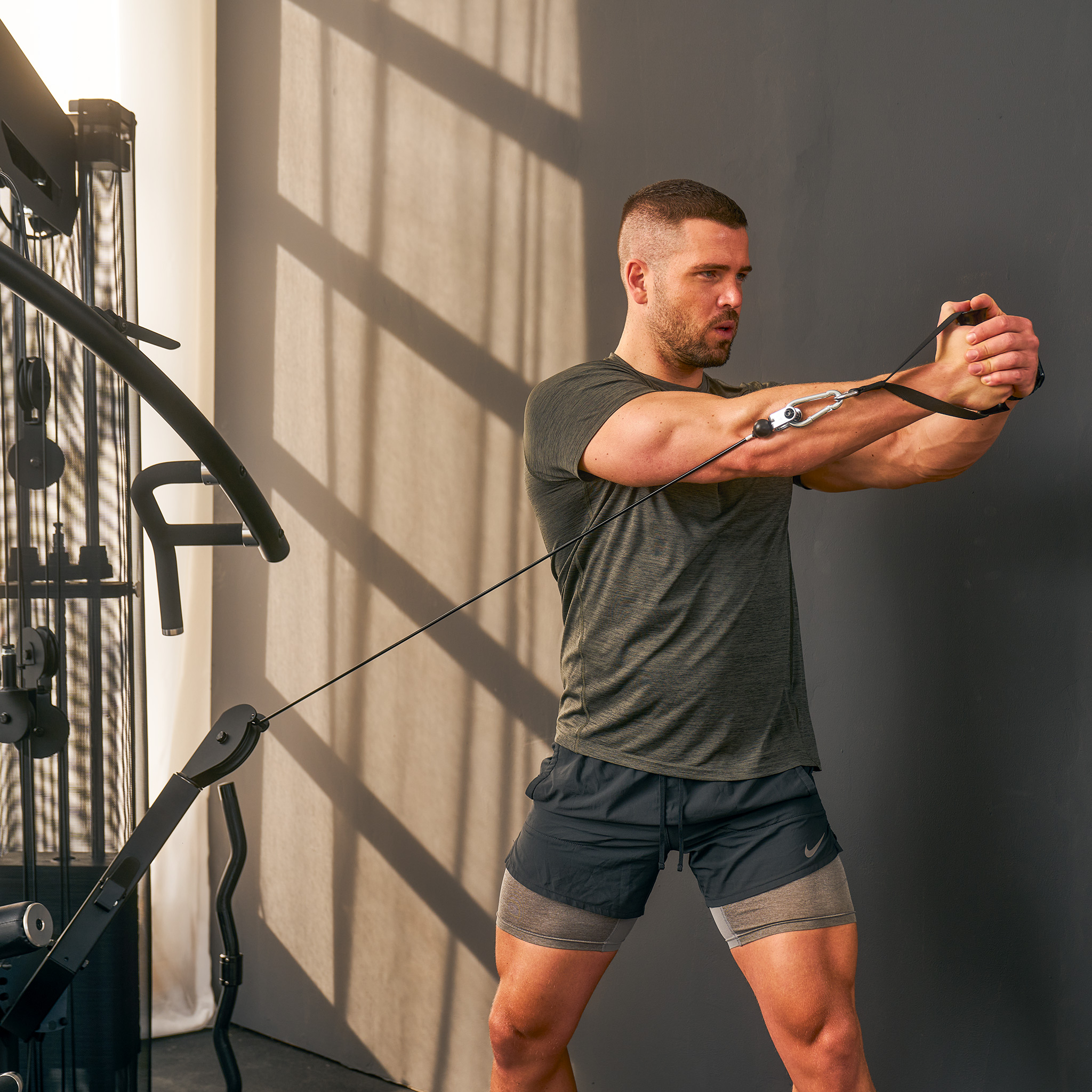 Ein Mann benutzt eine Kabelmaschine im Fitnessstudio und zieht Griffe zu seiner Brust. Er trägt ein dunkelgrünes Shirt.