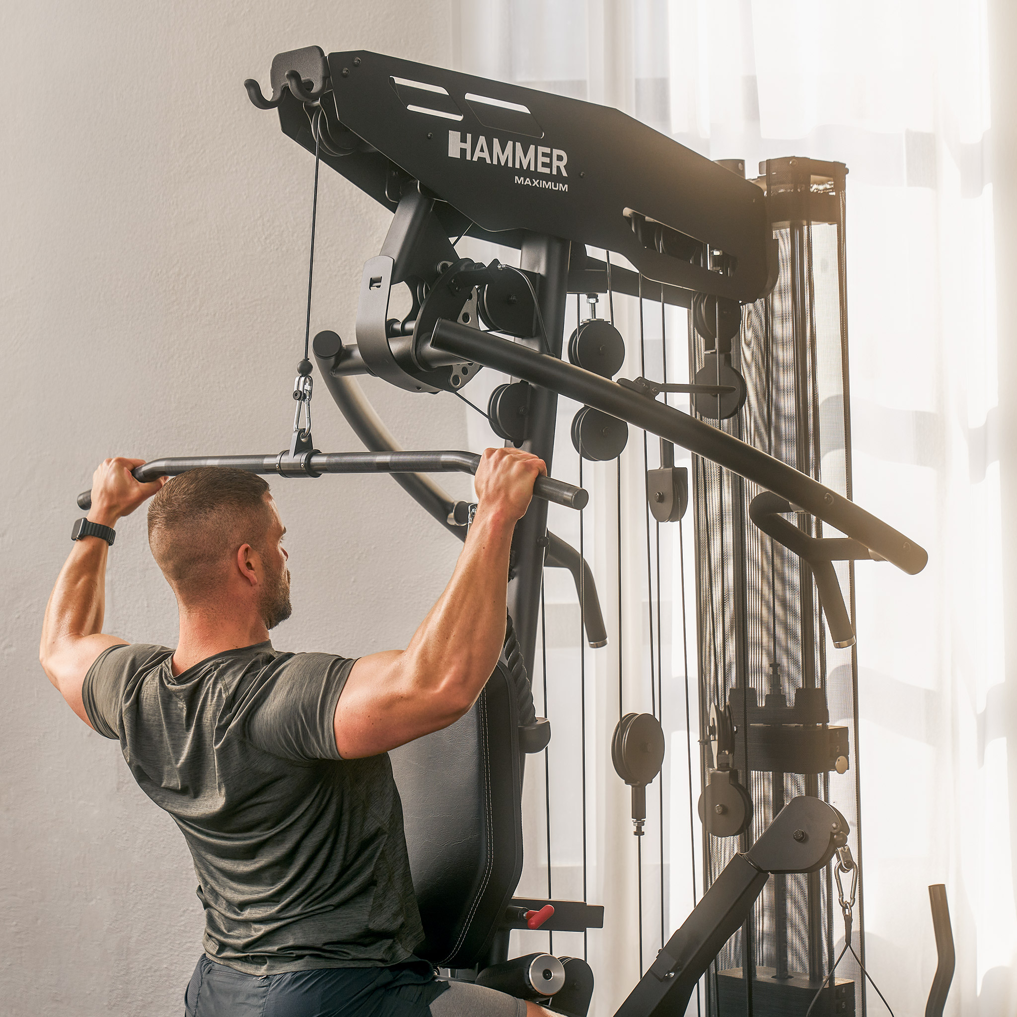 Mann benutzt schwarze Hammer-Fitnessmaschine, zieht eine Stange herunter. Weiße Wand und Fenster im Hintergrund.