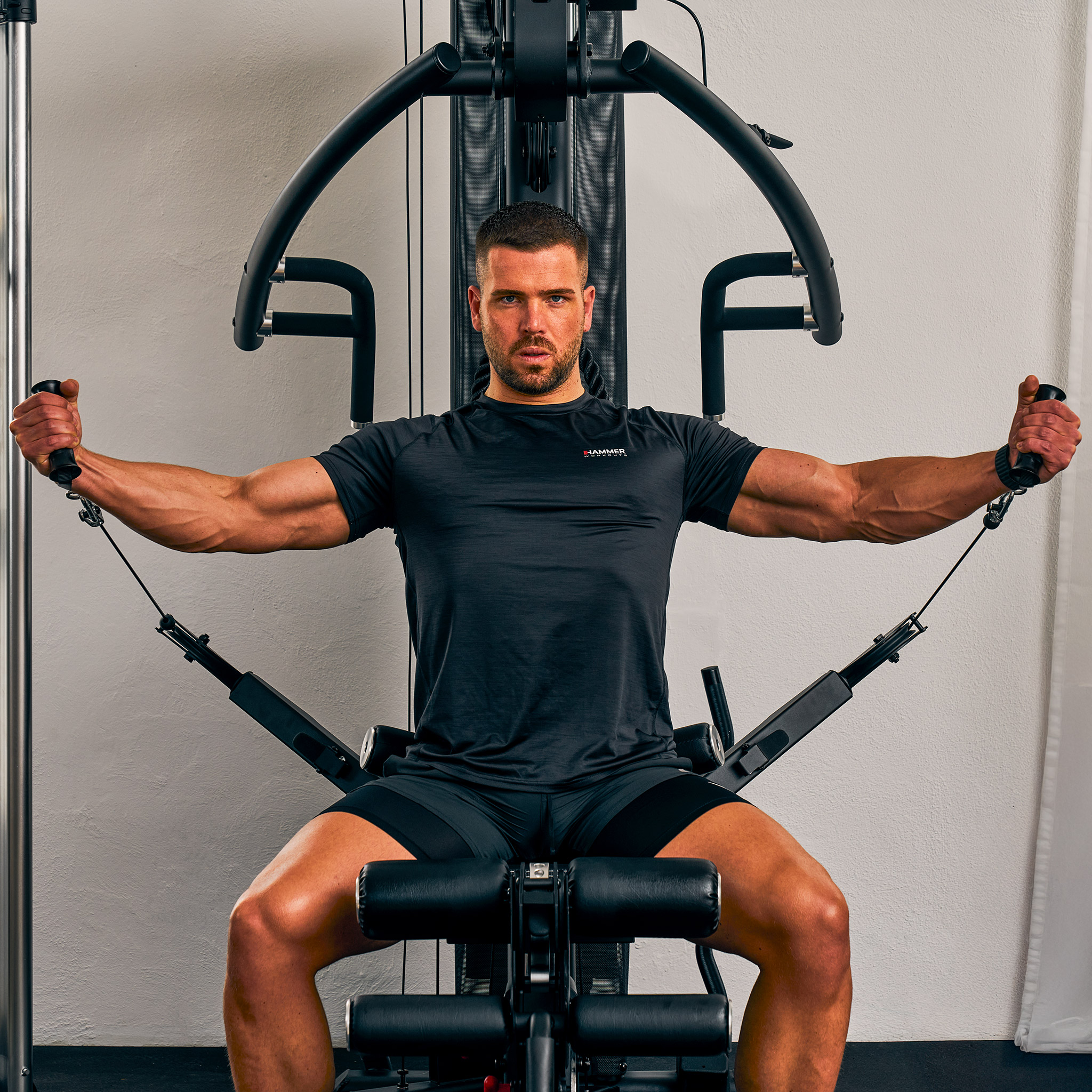 Ein Mann sitzt auf einem Fitnessgerät, Arme ausgestreckt, hält Griffe. Die Maschine ist schwarz mit einer silbernen Stange.