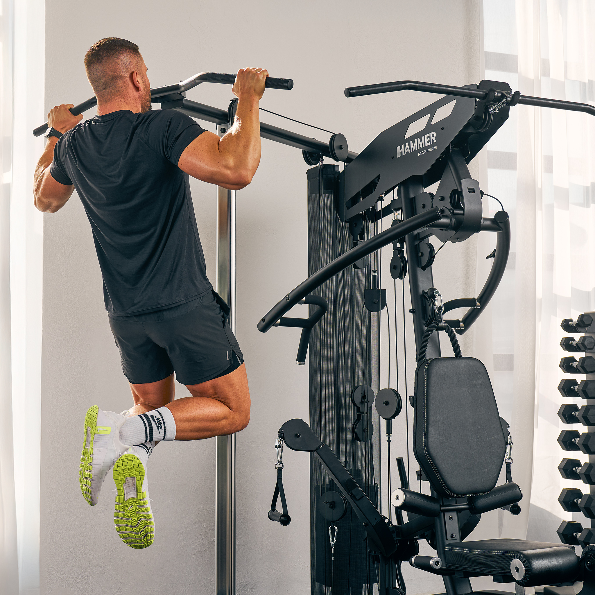 Mann, der Klimmzüge an einer schwarzen Trainingsmaschine macht. Er trägt schwarze Shorts und ein T-Shirt. Der Hintergrund ist ein weißer Raum.