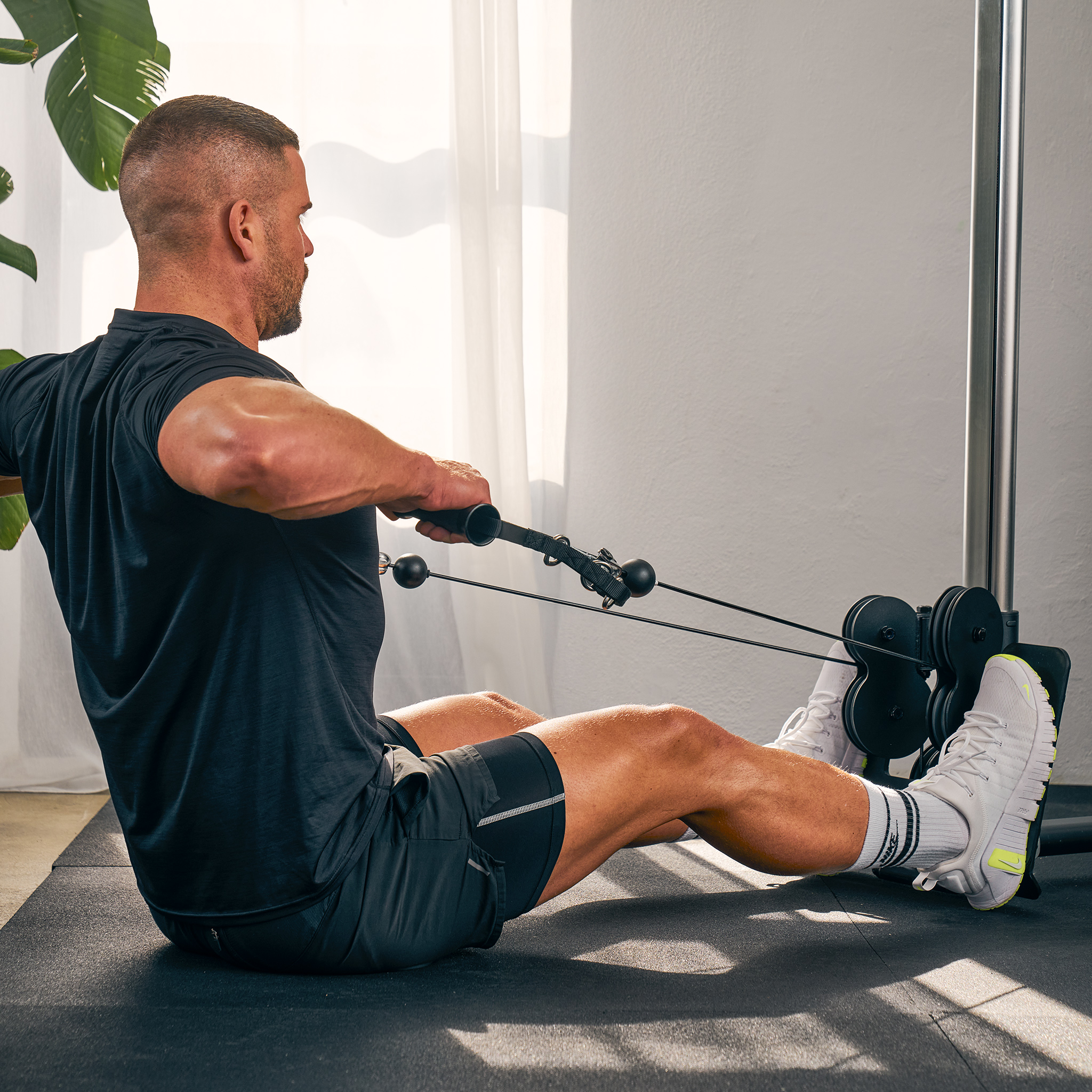 Ein Mann rudert auf einem Fitnessgerät, nach links blickend, mit dem Rücken zur Kamera, in einem Fitnessstudio, mit einer Pflanze sichtbar.